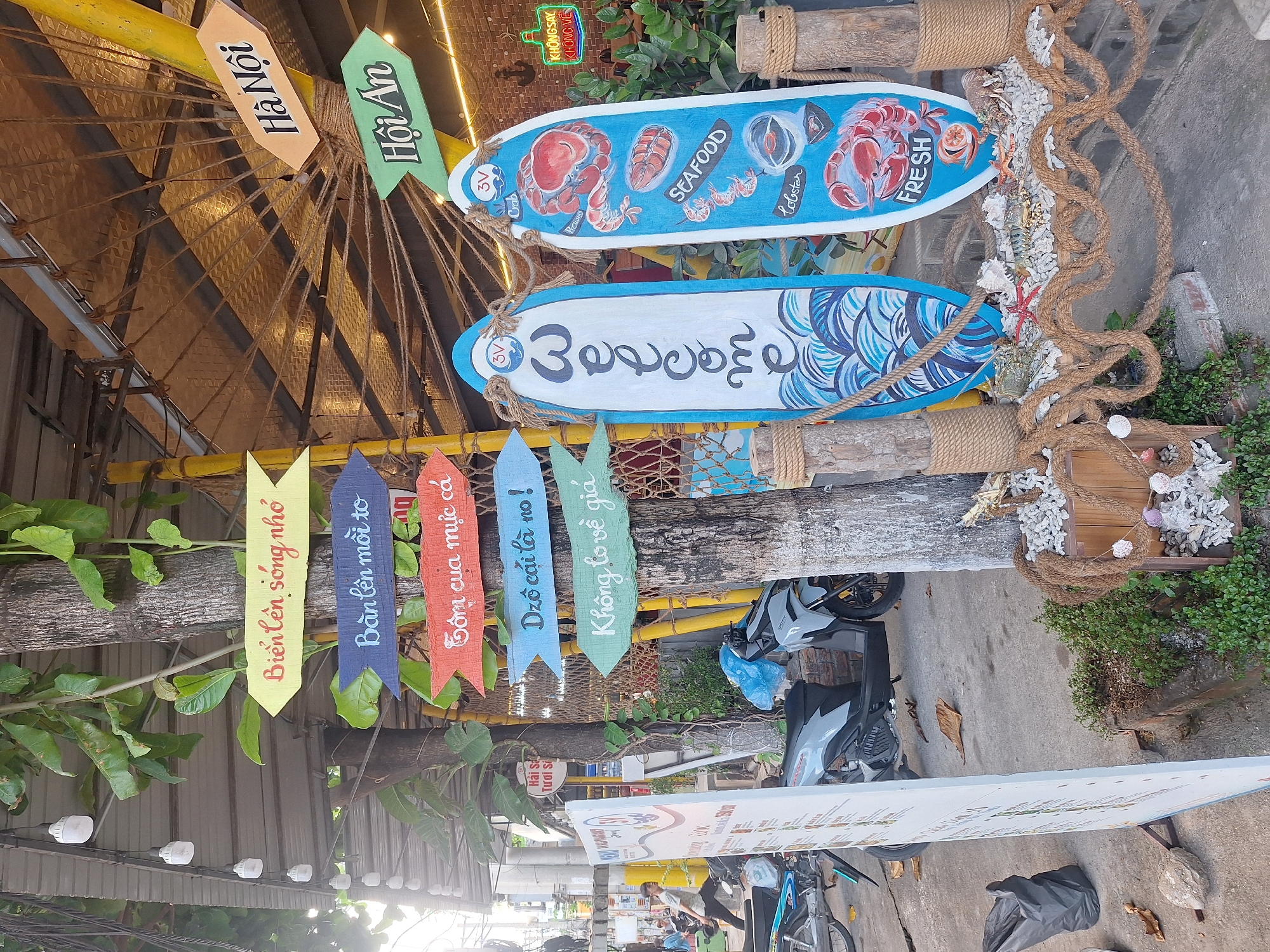 The image captures a street-level view in Nha Trang, Vietnam, featuring a cluster of directional signs attached to a tree and two surfboards used as decorative displays. The signs are colorful and point in various directions, with text in Vietnamese: "Biển lên sóng nhớ", "Bạn lên mỗi to", "Gom của mực cá", "Dzô cái là no!", and "Không lo về giá". Additionally, there are signs pointing towards "Hà Nội" and "Hội An".
The surfboards, standing upright and tied with rope, are painted with nautical themes. One surfboard has the word "Welcome" written vertically in white against a blue, wave-like pattern. The other surfboard displays illustrations of seafood, including crab, shrimp, and lobster, with the word "SEAFOOD" and "FRESH" prominently featured.
In the background, a building with a corrugated metal roof is visible, along with some greenery. To the left, a large white menu board stands near the street, and several motorcycles are parked nearby. A black trash bag lies on the ground, and fallen leaves are scattered on the pavement. The overall scene suggests an outdoor establishment, possibly a restaurant or cafe, located in a bustling urban environment.