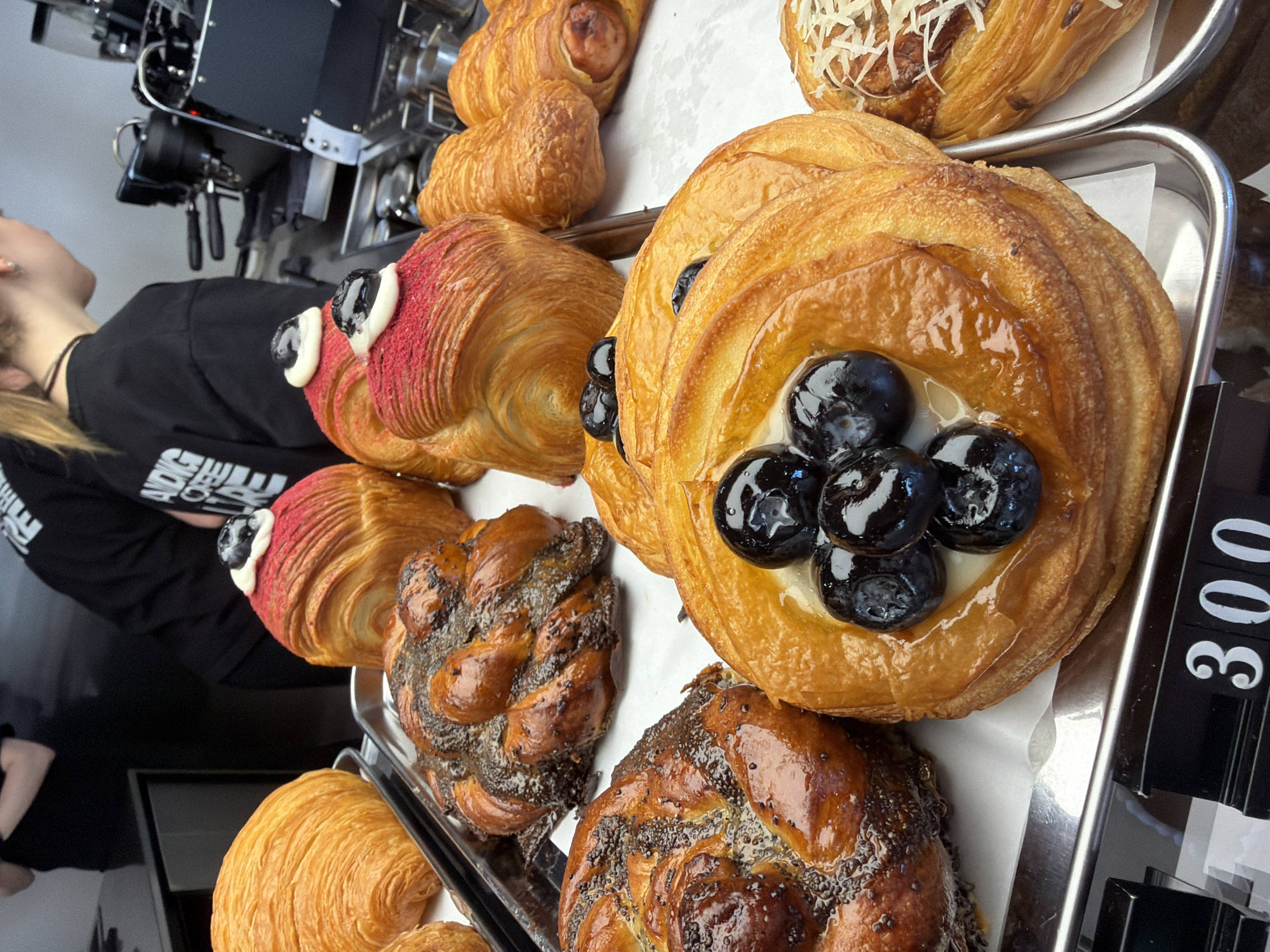 A close-up shot shows a tray of pastries in Izhevsk, Russia. The pastries include croissants with red icing and white dollops, twisted pastries with poppy seeds, a blueberry-filled danish, and a pastry with shredded cheese. A person wearing a black shirt is visible in the background. The price tag reads "300". A coffee machine is also visible in the background.