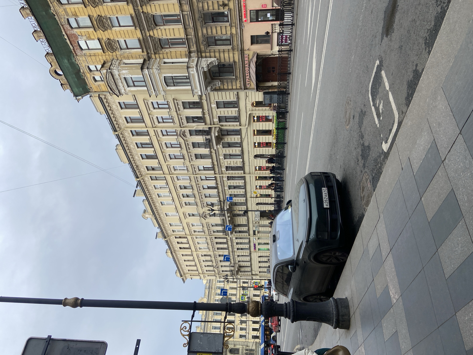 The image captures a street scene in Saint Petersburg, Russia. A dark green car is parked on the sidewalk, its license plate visible. A street lamp with ornate detailing stands on the sidewalk. In the background, a multi-story building with a yellow facade and numerous windows dominates the view. The street is marked with white lines and a bicycle symbol. Pedestrians can be seen on the opposite side of the street. Several street signs are visible. The sky is overcast.