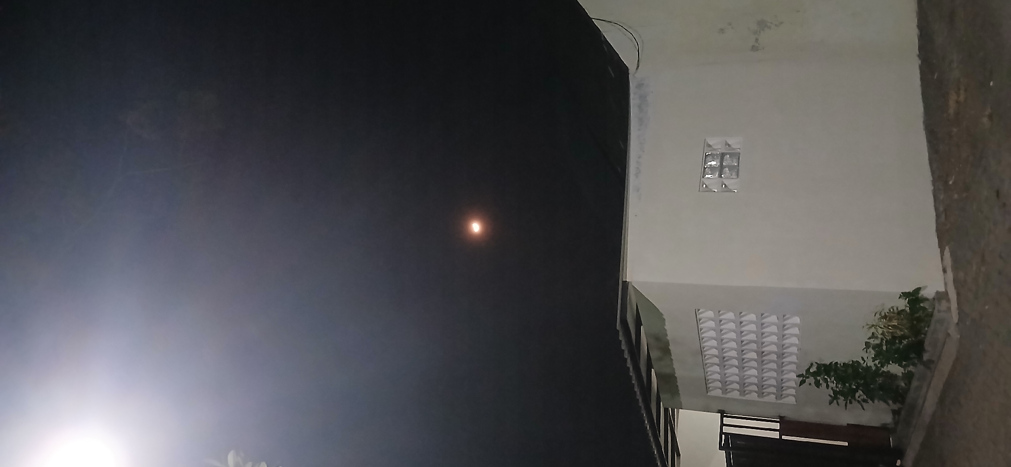 The image captures a night scene in Demangan, Indonesia. The sky is dark, with a bright, round object, possibly the moon or a strong light source, visible in the upper central portion. The lower half of the image features a light-colored building with a sloped roof supported by dark beams. The building has a decorative lattice-like pattern on its wall, and a small window made of glass blocks. A dark gate is present on the left side of the building, and a small plant is growing in front of it. The ground appears to be paved with gravel or small stones. The lighting suggests an artificial light source is illuminating the building.