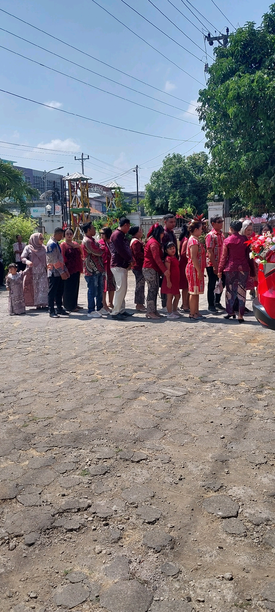 In Keling, Indonesia, a group of individuals is lined up outdoors on a cracked asphalt surface. The people are dressed in traditional Indonesian attire. Some are wearing long-sleeved shirts and pants, while others have on dresses or skirts. Many of the garments feature vibrant colors and intricate patterns. A few of the women have their heads covered with headscarves. Children are also present in the line. In the background, there are buildings, trees, and utility poles with electrical wires. A red vehicle is partially visible on the right side of the image. The sky above is clear and blue with a few scattered clouds. The overall scene suggests a formal gathering or event taking place during the daytime.