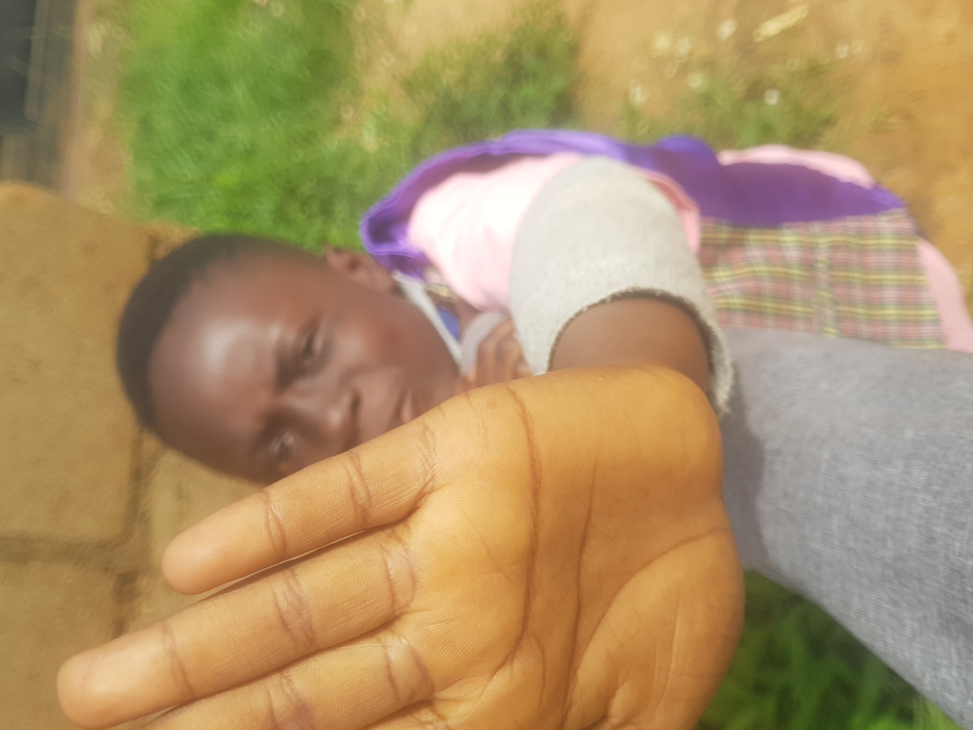 A close-up shot captures a child's left hand held up to the camera, obscuring a significant portion of their face. The child, with dark skin and hair, wears a pink shirt and a plaid garment layered underneath with a purple sash draped over their shoulder. Their expression is partially visible, showing a furrowed brow and a slightly open mouth. The background is blurred, suggesting an outdoor setting with green foliage and a sandy or dirt ground. The overall lighting is bright, indicating daytime.