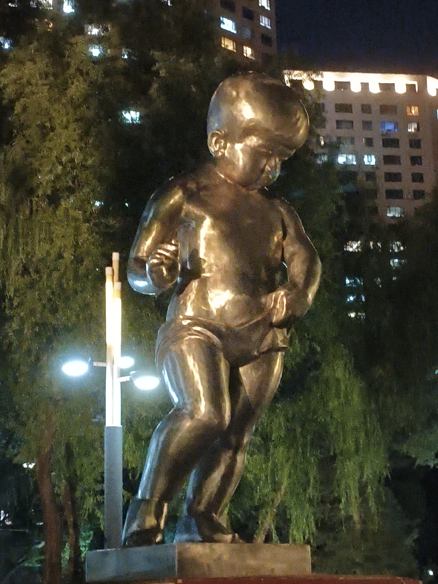 The image shows a bronze statue of a toddler in Dongxing Street, China, at night. The statue is located in front of a building with illuminated windows and is surrounded by trees. A lamppost with multiple lights is visible to the left of the statue. The statue depicts a child standing with its legs slightly apart and its arms bent, holding onto its shorts. The lighting in the scene is dim, with the primary light sources being the building's windows and the lamppost.