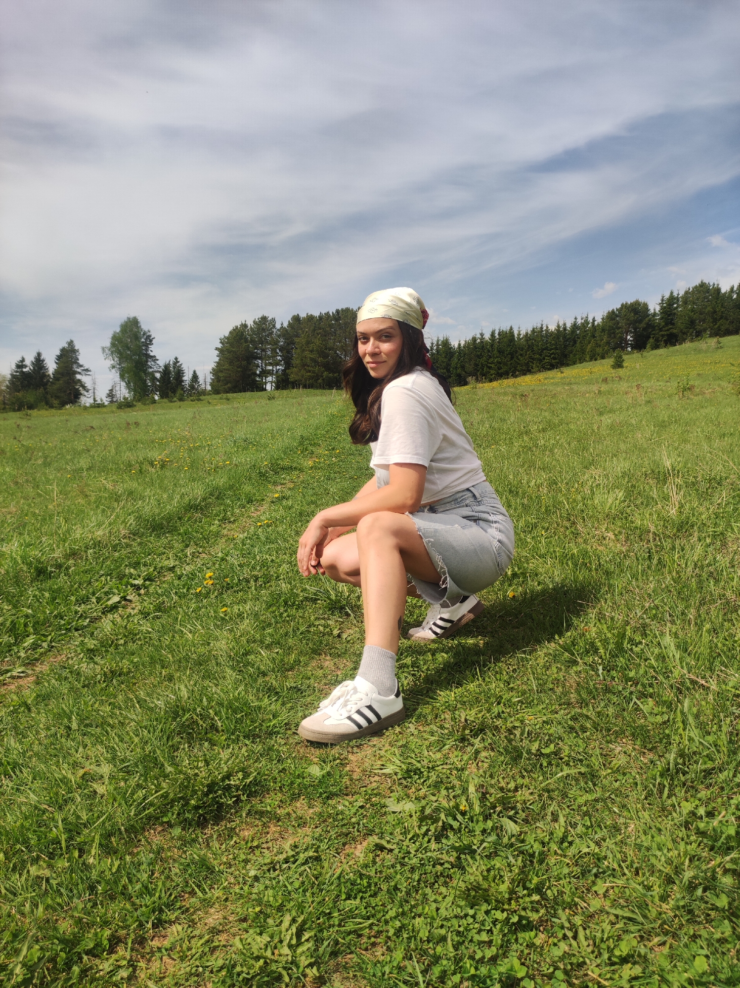 A young woman is squatting in a grassy field. She wears a white t-shirt, denim shorts, grey socks, and white Adidas sneakers with black stripes. A patterned bandana covers her head. The background shows a green meadow sloping up to a line of trees under a partly cloudy sky. Based on the location, the setting is likely the region surrounding Фоки, Russia, known for its natural landscapes.