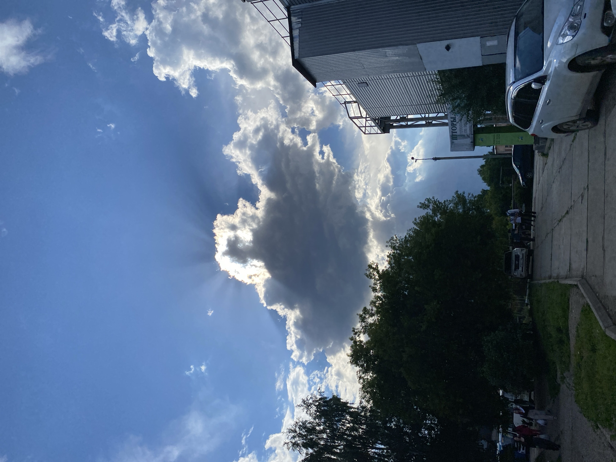 The image depicts a cloudy sky with sunbeams radiating from behind a large cloud, creating a dramatic effect. In the foreground, there is a paved area with a sidewalk, some grass, and trees. A silver SUV is parked on the right side of the image. To the left, a few people are walking on the sidewalk, and several cars are parked. A tall, industrial-looking building with a corrugated metal exterior is visible on the right. A sign with Cyrillic text, possibly for curtains or blinds, is mounted on a pole in the middle ground. The scene appears to be an outdoor urban environment in Votkinsk, Russia.