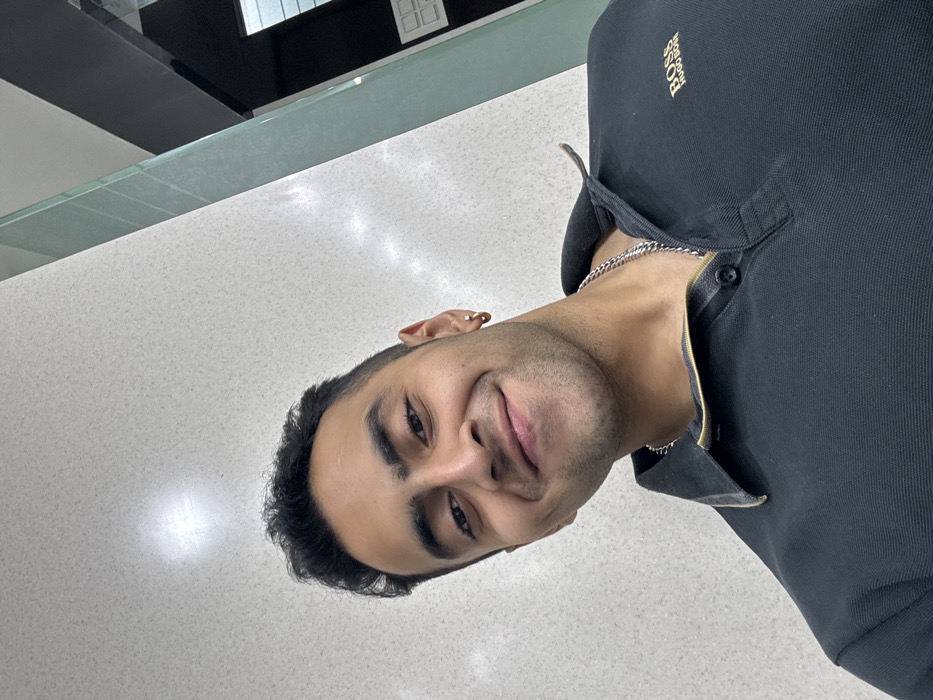 A close-up, upward-angled portrait depicts an adult male of South Asian ethnicity within an indoor setting in Kolkata, India. The subject presents a smiling expression, displaying dimples and light facial stubble. He has dark, short-styled hair. He wears a dark polo shirt featuring a gold-toned collar trim and a gold "BOSS HUGO BOSS" logo on the left chest. A silver-colored chain necklace is visible, alongside a gold-colored hoop earring in his left ear and a small stud earring in his right ear. The background consists of a light-colored, speckled wall reflecting overhead lighting on the left and center, adjoined by a dark, reflective vertical panel on the right.
