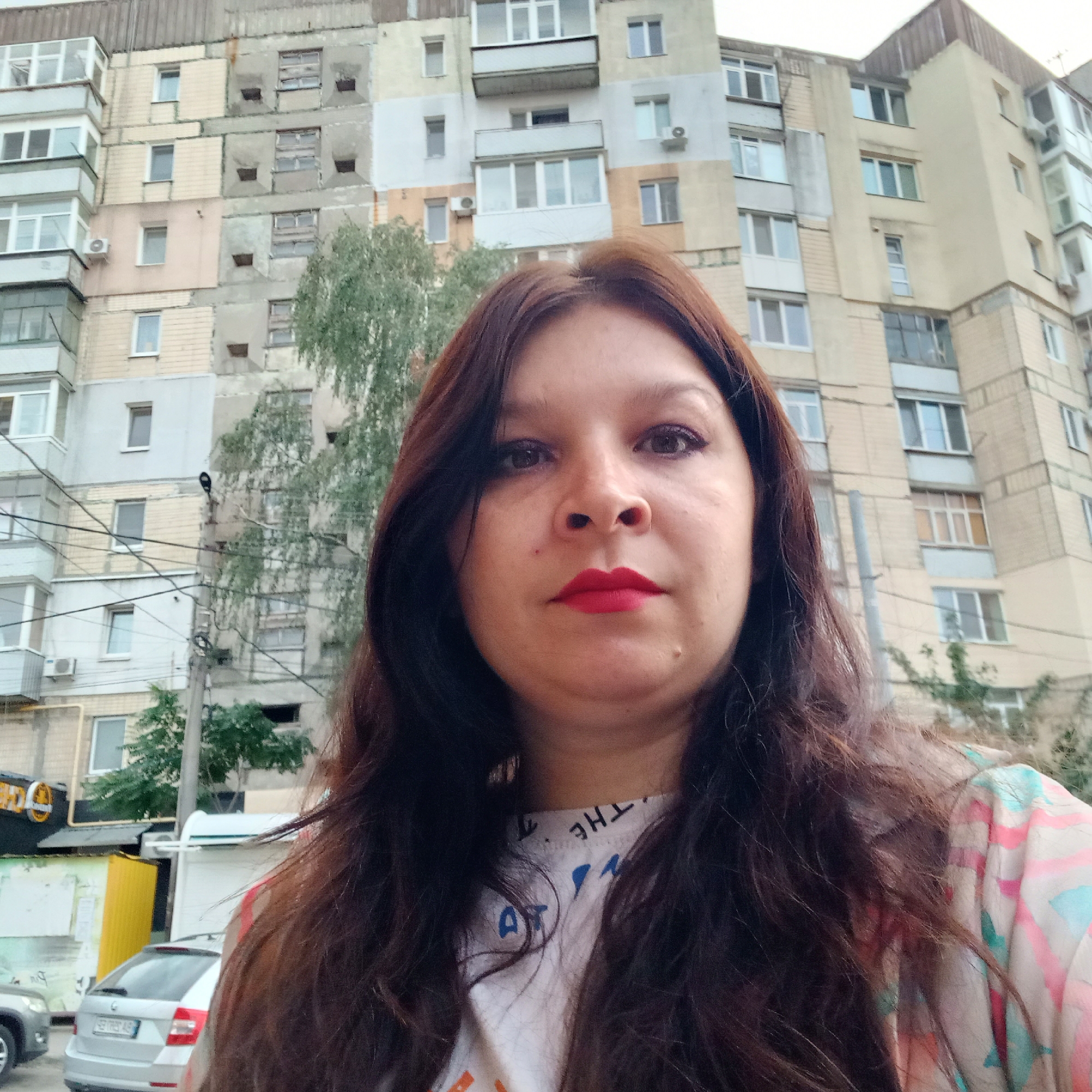 A selfie of a woman with long, dark brown hair and red lipstick is taken from the chest up. She is standing in front of a weathered apartment building in Kropyvnytskyi, Ukraine. The building has a mix of beige, grey, and light pink panels, with numerous windows and balconies. A large tree with green foliage is visible to the left of the woman. In the background, a white car and a yellow structure are also visible. The woman is wearing a white t-shirt with some text and blue and orange lettering on it. Her clothing has a colorful geometric pattern on it. The sky is overcast.