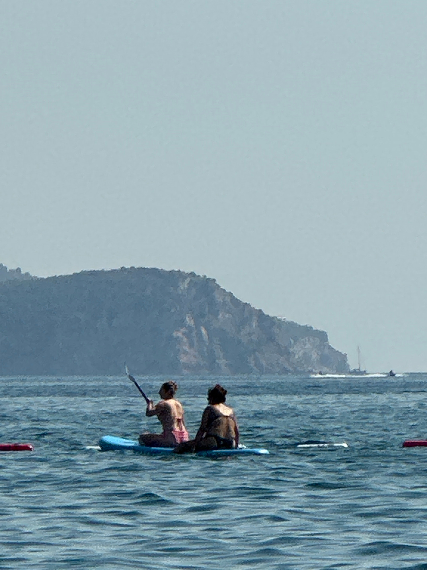 Two individuals are sitting on a paddleboard in the water, with a mountainous coastline in the background. The person in the front is wearing a pink bikini and is holding a paddle. The person behind them is wearing a brown swimsuit. The water is blue and there are ripples on the surface. The sky is clear and hazy. The location is the city of Boreti, Montenegro.