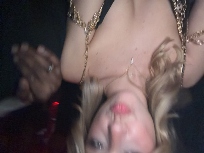 An image, blurry and underexposed, depicts a blonde female with light skin, positioned centrally and to the left, facing the camera with puckered lips. She wears a black garment adorned with intricate gold-colored chain-link shoulder and arm jewelry, along with a thin gold necklace featuring a small, light-colored pendant. To her right, partially visible against a dark background, is a dark-skinned hand wearing a silver-colored ring on the ring finger. A faint red light source is discernible in the upper right background. The scene is set in a dimly lit interior environment, likely a social venue. The precise location in the city of Paris, France, or any other specific city, cannot be determined from the visual information provided.