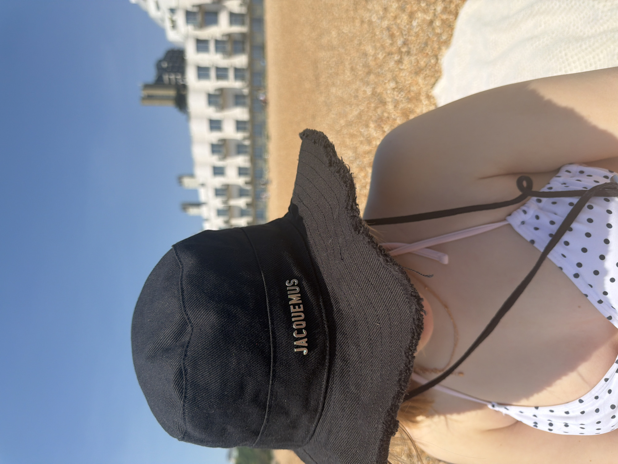 A daytime eye-level shot captures a person wearing a black "JACQUEMUS" branded hat and a white and black polka dot bikini top. The person is on a beach with light brown sand and a white object behind them, possibly a towel. In the background, a multi-story white building with multiple windows is visible, suggesting an urban beach scene in Folkestone and Hythe District, United Kingdom. The sky is clear and blue.