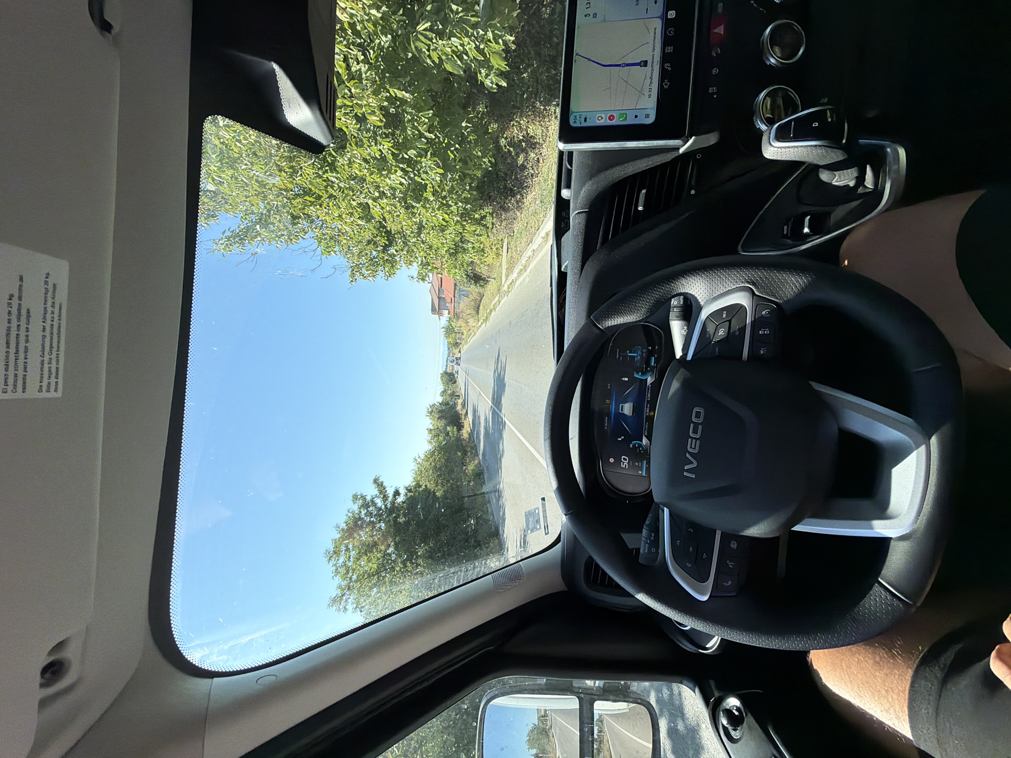 The image shows the interior of a vehicle driving on a road. The perspective is from the driver's seat, looking forward through the windshield. The dashboard features a steering wheel with the "IVECO" logo, a digital instrument cluster displaying a speed of 50, and a touchscreen navigation system showing a map. Sunlight streams through the windshield and the driver's side window, illuminating the road ahead and the surrounding lush green trees. The road appears to be paved and is bordered by vegetation on both sides. The sky is clear and blue. The vehicle is likely in a rural area or on the outskirts of a town, given the presence of trees and what appear to be houses in the distance on the right side of the road. There is a person's legs and upper thighs visible in the lower part of the image, indicating the driver or a passenger. The location is specified as Trapishte, Bulgaria, implying the scene is taking place within this geographical area.