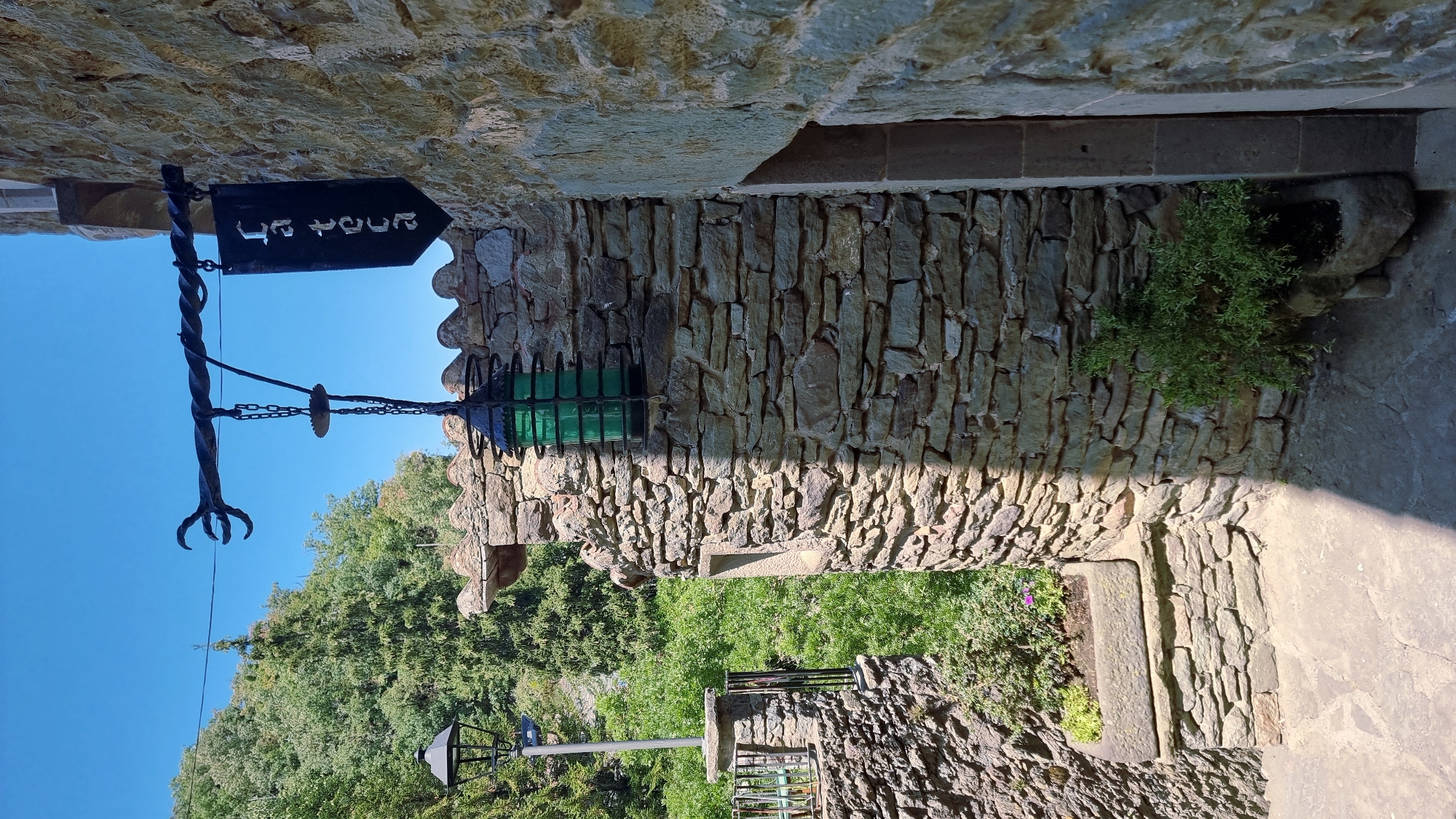 This image depicts a rustic stone building with a hanging sign and lantern. The sign, a black banner with white lettering, reads "La teca". A green glass lantern hangs from a wrought-iron fixture, which also supports the sign. The building is constructed of rough-hewn stones, characteristic of the medieval architecture found in Rupit i Pruit, Spain, a village renowned for its well-preserved Catalan Romanesque and Gothic buildings. The background features lush green trees and a clear blue sky. A lamppost stands to the left, and a stone planter with greenery is visible in the foreground. The scene suggests a historical setting, likely a shop or restaurant, given the signage.