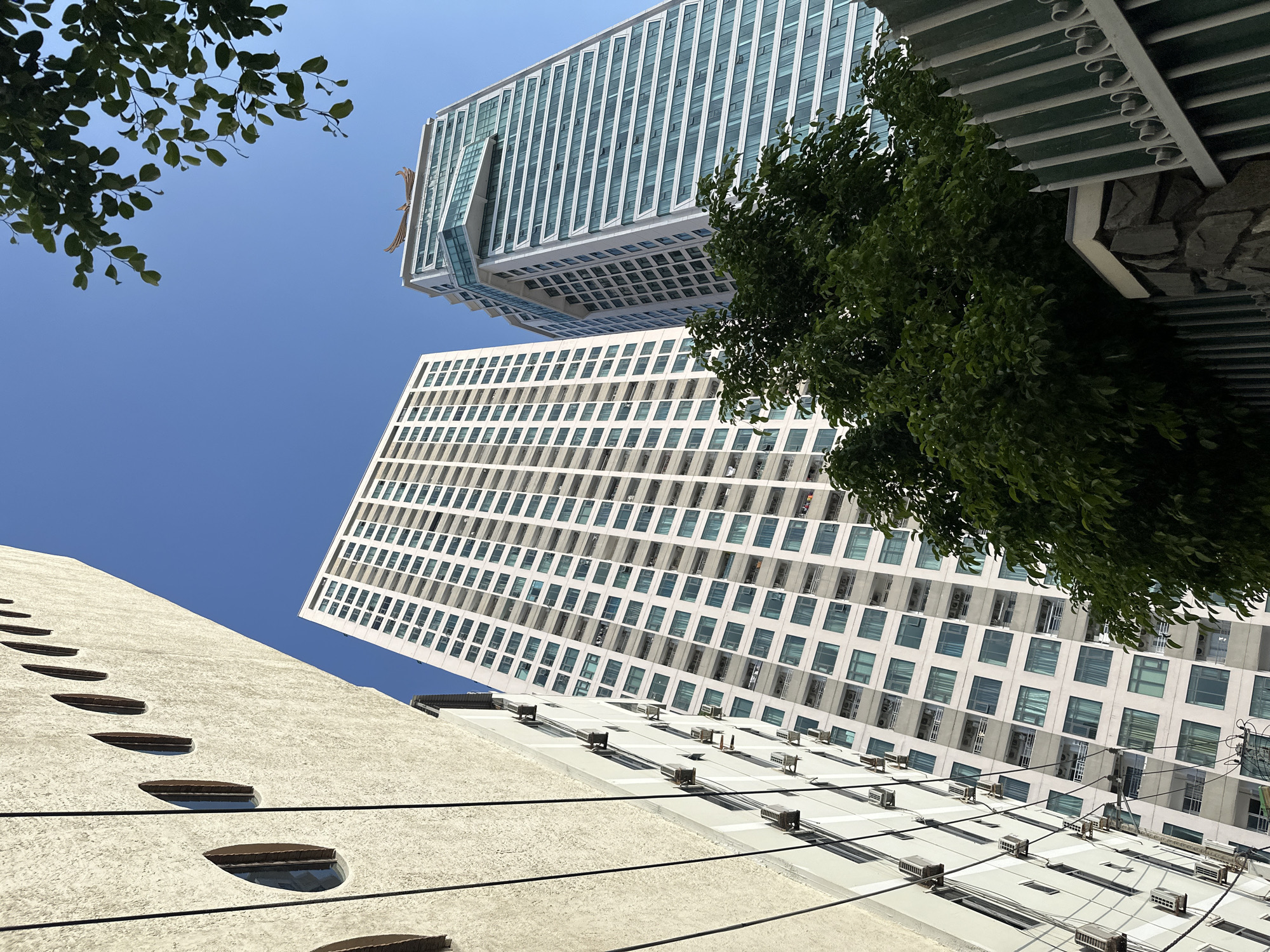 The image shows an upward view of buildings in Bac Nha Trang Ward, Vietnam, under a clear blue sky. From the bottom left, a beige building with teardrop-shaped windows is partially visible. Above it is a tall, white building with many windows arranged in a grid pattern, and numerous air conditioning units affixed to its exterior. To the right of the white building, a green tree partially obstructs the view. Another building, with a unique design featuring a cantilevered section, is at the top center of the image. Green tree leaves are visible at the top left, with an architectural structure with a green and white color scheme at the top right of the scene. Electrical wires intersect the lower portion of the image.