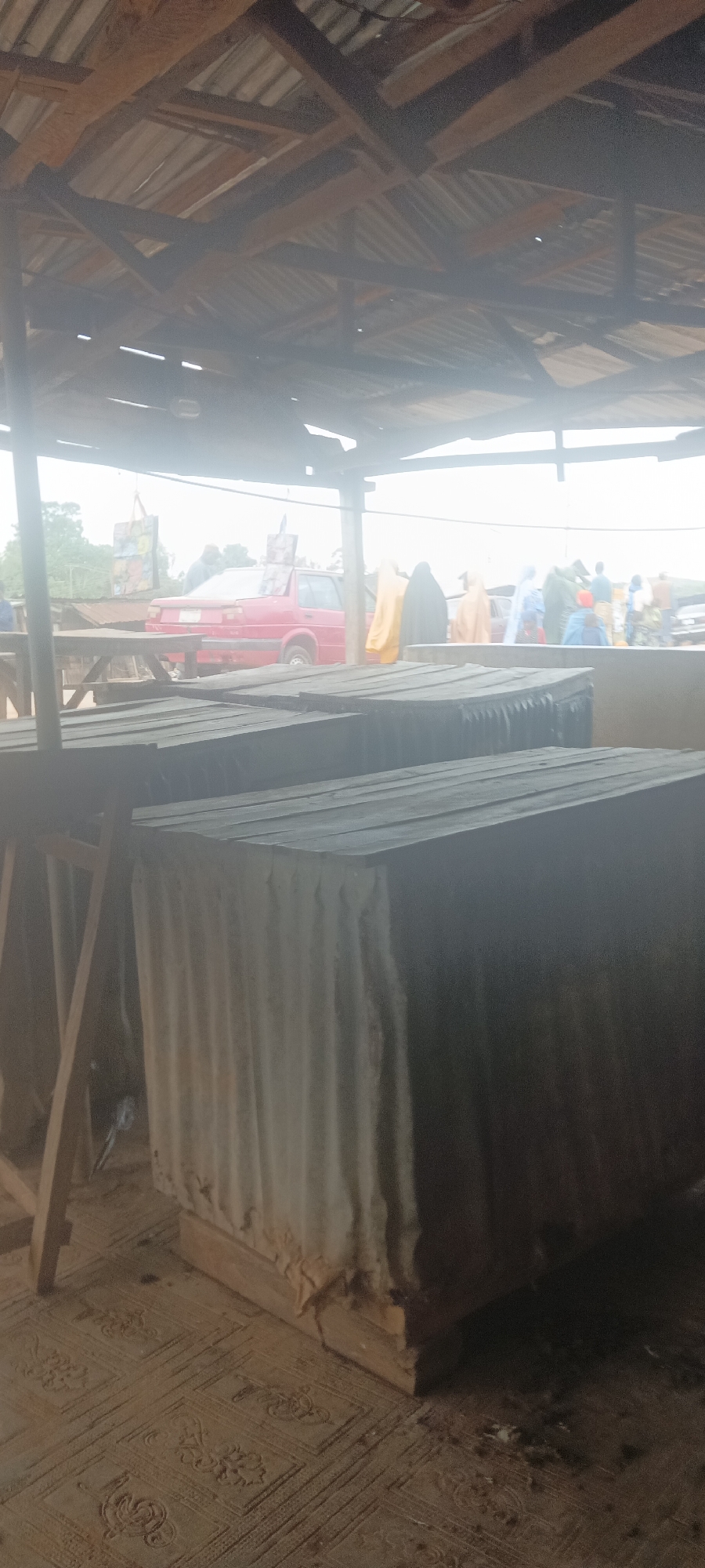 The image depicts a scene at Kassa Market, Nigeria. Underneath a corrugated metal roof, there are several rectangular structures with corrugated metal sides, likely stalls or storage areas. In the background, there are people and a red car visible through the open sides of the market structure. Some people are wearing garments with different colors. The foreground shows the floor, which appears to be patterned. There is a wooden support structure visible in the left foreground. The overall lighting is dim, possibly due to the roof cover and weather conditions.