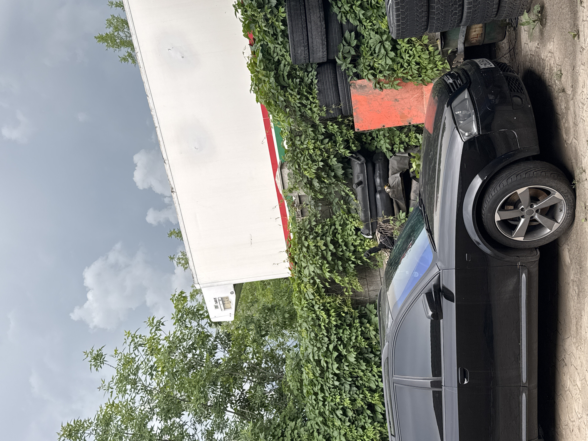 A black Audi A6 Avant is parked on a cobblestone driveway in front of a white semi-trailer truck. The truck is partially obscured by dense green foliage and a stacked pile of tires. The truck has a red and green stripe along its side. The sky above is cloudy and grey. The overall scene appears to be an outdoor storage or industrial area.