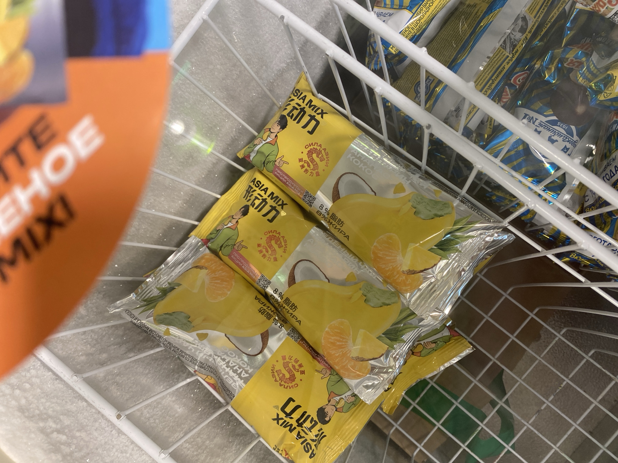 A close-up, slightly tilted overhead shot shows several yellow ice cream wrappers inside a white wire freezer basket. The wrappers depict fruit imagery, including oranges and possibly mangoes, along with cartoon characters. Some wrappers also feature Asian script. The background is blurred, revealing a frosty freezer interior. The image appears to have been taken in a commercial setting, likely a store, and is consistent with being in Lomonosov, Russia, given the presence of Russian text on some of the packaging.