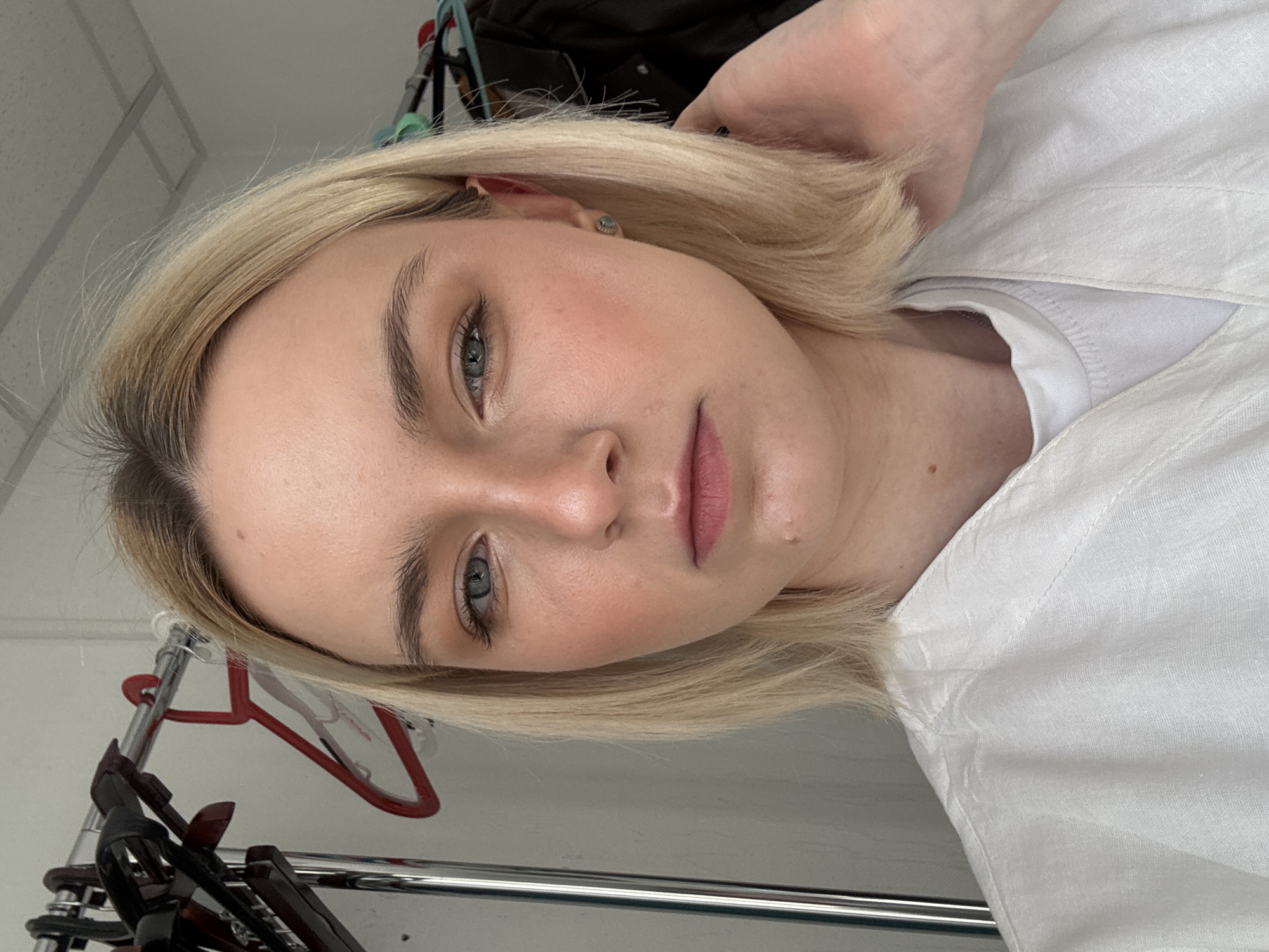 A close-up, medium shot depicts a fair-skinned woman with blonde, shoulder-length hair, appearing to be in a casual setting. She has light blue eyes and a neutral facial expression. She is wearing a white, collared shirt, partially revealing a white undershirt. The background reveals a clothing rack with hangers. The location cannot be determined from the image.