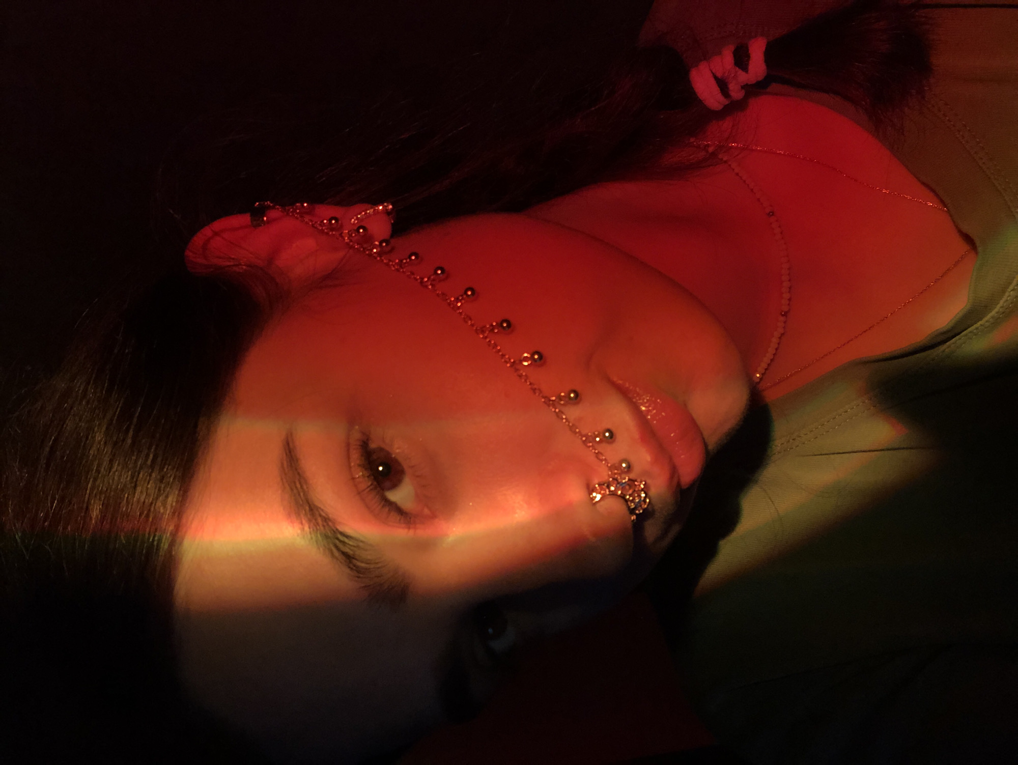 In this close-up, dimly lit, medium shot portrait, a young person with dark hair and brown eyes is featured against a dark background. The subject is adorned with elaborate gold jewelry. A nose ring with intricate detailing and dangling charms is visible, connected by a delicate chain to an earring that extends upwards and attaches to the ear. A faint rainbow-colored light effect casts a stripe across the subject's face, highlighting their left eye and cheekbone. The subject wears a green garment. The image does not provide any information about the location in the city of Noviy, Russia.