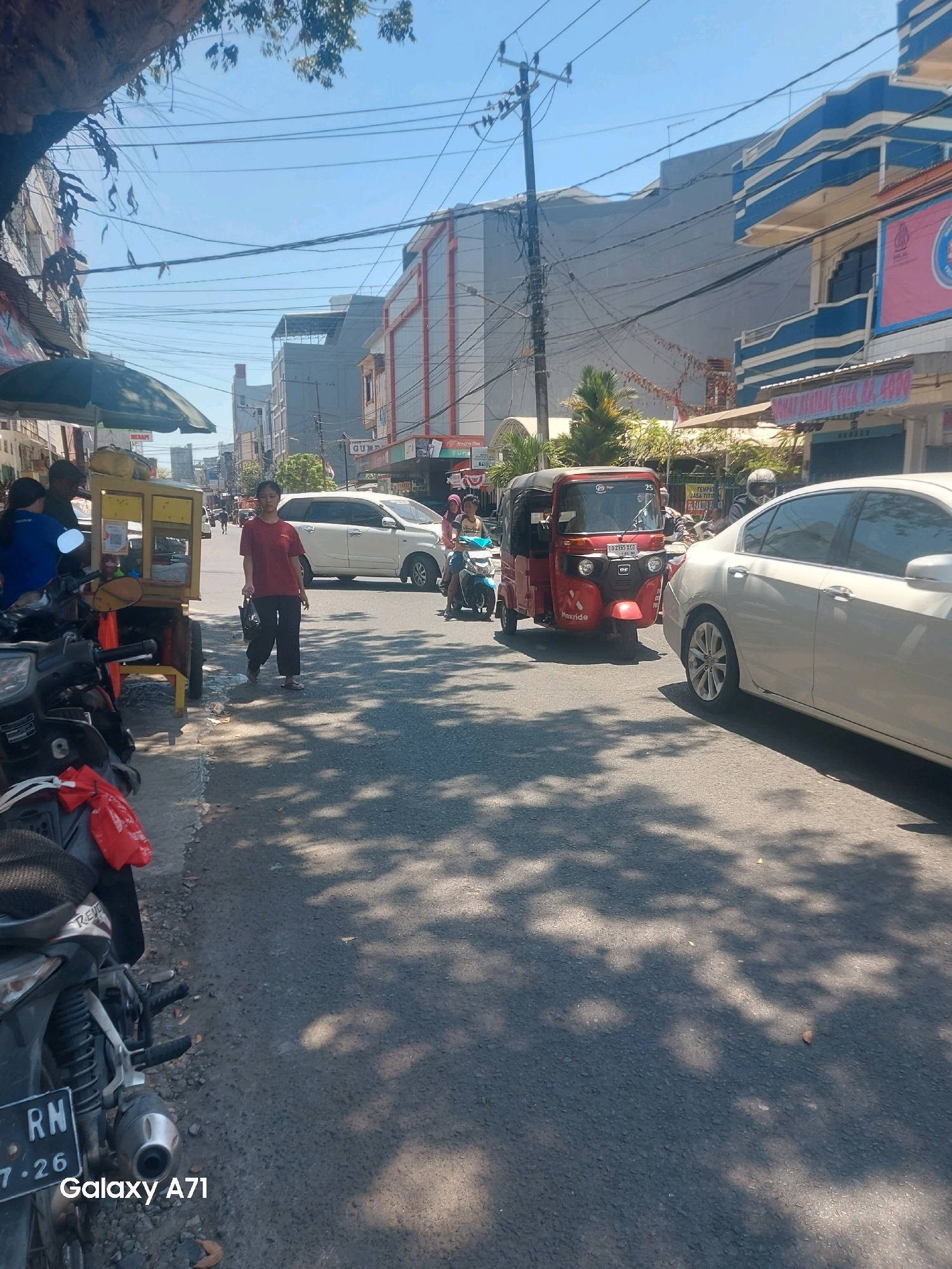 On a sunny day in Makassar, Indonesia, a street scene unfolds under a canopy of power lines and tree branches. The asphalt road is shaded by dappled sunlight, with fallen leaves scattered across its surface. To the left, a sidewalk is lined with a food cart adorned with a green umbrella, and several motorcycles are parked along the curb, one with a red plastic bag tied to its handlebars. A woman in a red shirt and black pants walks away from the cart, carrying a dark bag. Beyond her, a white SUV is visible on the road. In the center, a red three-wheeled vehicle, identified as a "Haxride," travels forward. Further down the street, buildings with blue and white architectural details and various signage indicate a commercial area. The sky is a clear, bright blue.