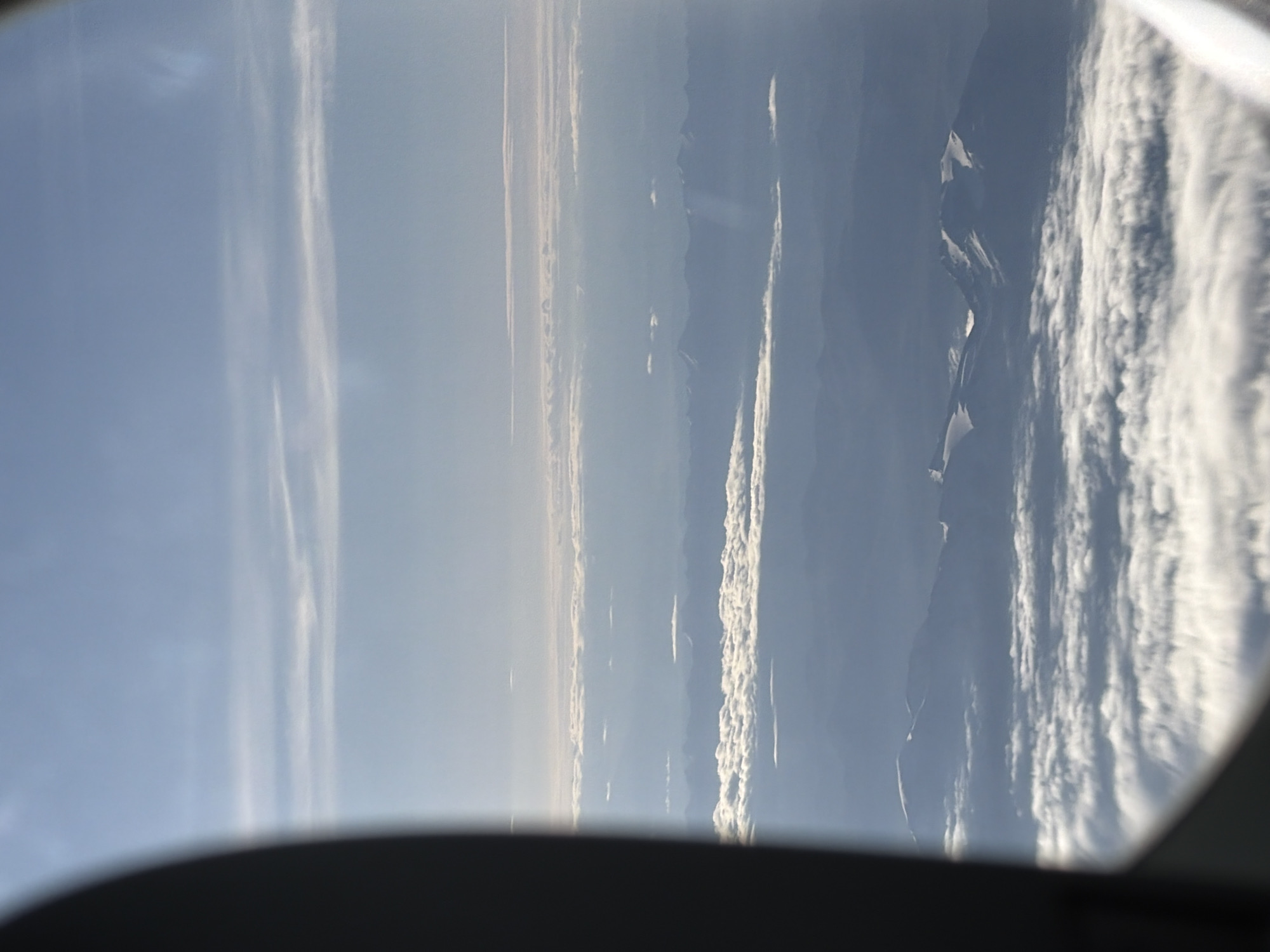 The image shows a view from an airplane window, looking down on layers of clouds and mountains. The left side of the frame is dominated by the dark, curved edge of the airplane's window frame. Above the clouds, the sky is a pale blue with wispy white clouds. Below the sky, several layers of clouds are visible, some appearing flat and spread out, while others are more textured and billowy. In the distance, snow-capped mountains are visible, appearing hazy and layered behind the clouds. The lighting suggests it is daytime, possibly early morning or late afternoon, with a soft, diffused light. There are no people, specific landmarks, or any indication of the city of Hiranagar, India. The scene is entirely natural and aerial.