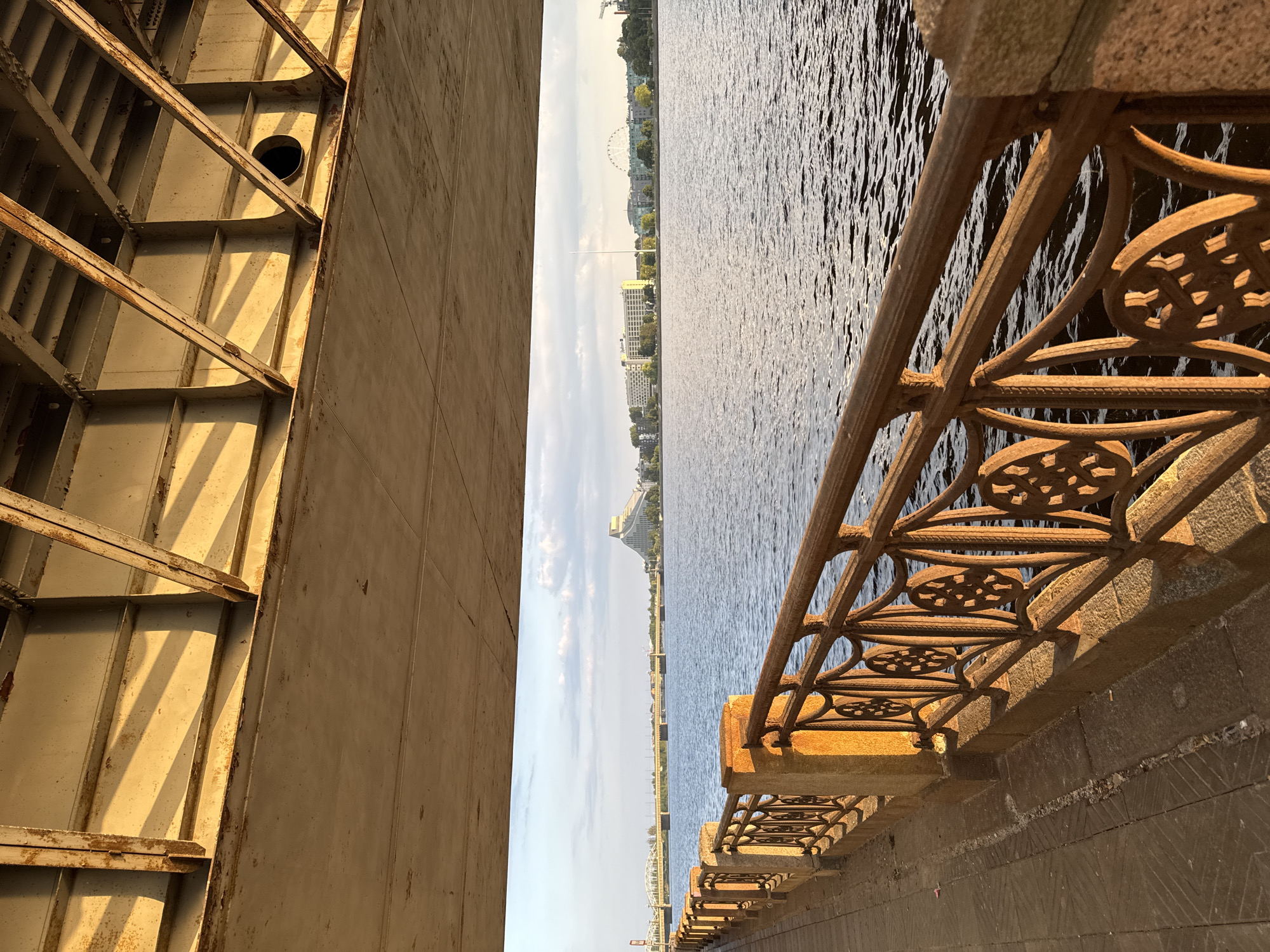 The image captures a view of the Daugava River in Riga, Latvia, as seen from beneath a bridge, likely the Vanšu Bridge. The foreground is dominated by the weathered concrete underside of the bridge, with exposed metal girders showing signs of rust. Below the bridge's structure, a promenade runs alongside the river, bordered by an ornate, wrought-iron railing with decorative circular patterns. The railing and its stone supports are illuminated by the warm, golden light of the setting sun, casting long shadows.
Across the river, the cityscape of Riga unfolds. Notable landmarks visible include the distinctive triangular roof of the Latvian National Library on the left bank and a Ferris wheel on the right bank, suggesting the location is near the city center. Buildings line the riverfront, including a large hotel with a prominent "Radisson" sign. The Daugava River itself is calm, with gentle ripples on its surface reflecting the sky and the surrounding structures. The sky is a mix of blue with scattered white clouds. No people are visible in the scene. The overall impression is one of an urban waterfront scene bathed in the soft light of dusk, with elements of both industrial infrastructure and historic architecture.
