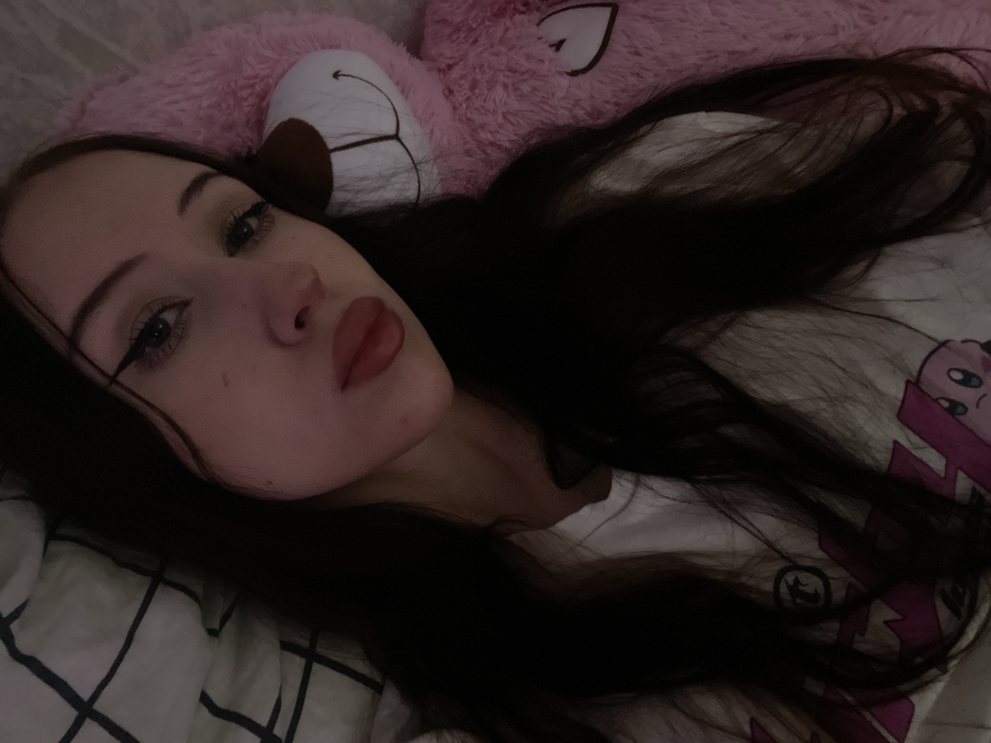 The image shows a young person with long, dark hair, lying down and partially obscured by a large pink teddy bear. The person is wearing a white t-shirt with a graphic print. The background includes a checkered blanket or comforter. The lighting is dim, suggesting an indoor setting, possibly a bedroom. There are no identifiable landmarks or features that would indicate a specific location in Votkinsk, Russia.