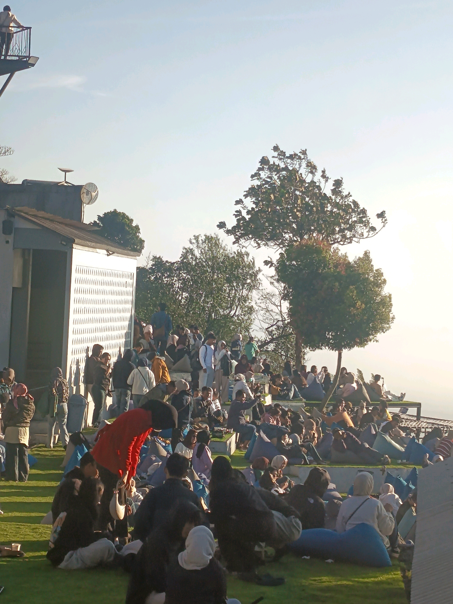 The image depicts an outdoor gathering of people in what appears to be a park or recreational area in Keditan, Indonesia. The setting is characterized by a large crowd seated on the grass and on tiered seating areas, many of whom are lounging in beanbag chairs. The environment is grassy, with several trees providing shade. In the background, a building with a distinct textured facade is visible, along with a balcony structure extending from a higher level. A person is standing on this balcony, looking out. The overall atmosphere seems relaxed, with many individuals engaged in casual conversation or simply enjoying the scenery. The sky is bright and clear, suggesting a pleasant day.