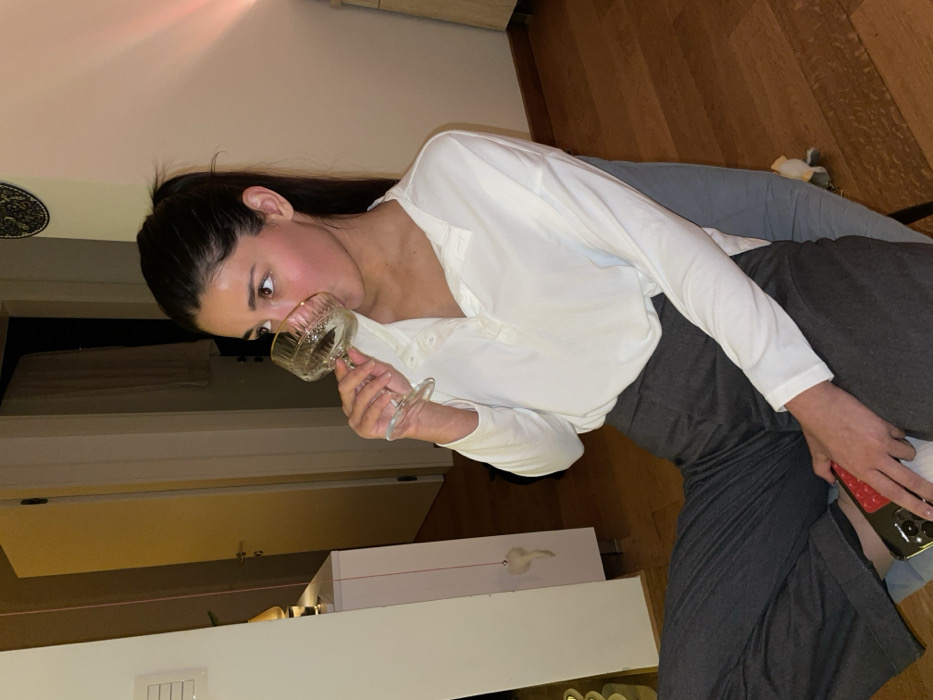A woman with dark hair in a high ponytail takes a sip from a stemmed glass. She wears a white, button-down shirt and grey pants while sitting on a light blue couch. Her eyes are wide and looking off to the left. A phone rests in her lap. The scene appears to be indoors, with a door visible on the left and wooden flooring on the right. A pink light is cast on the ceiling.