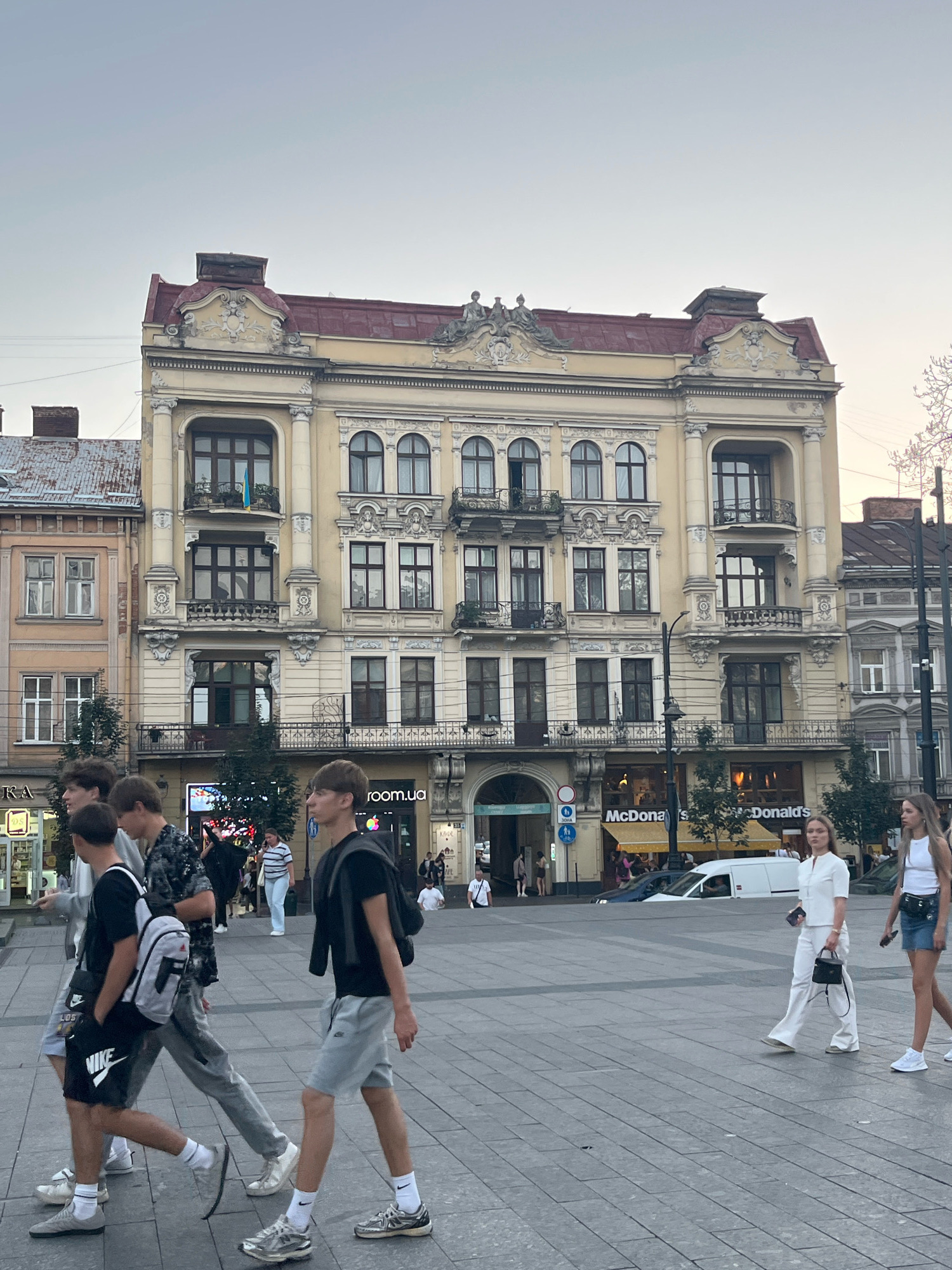 This image shows a street scene in Lviv, Ukraine. In the foreground, four young men are walking across a paved plaza. The man on the left is wearing a black t-shirt, black shorts with a white Nike logo, and white socks with grey sneakers. He has a grey backpack on his back. Next to him, a man in a grey t-shirt and grey pants is walking. To his right, a man in a black and white patterned shirt and grey pants is walking. The man on the far right is wearing a black t-shirt, grey shorts, white socks, and silver sneakers. He has a black backpack.
In the background, a large, ornate building with yellow facade and a red roof is visible. The building has multiple windows and balconies. Below the building, there are several businesses including McDonald's. A white van is parked in front of McDonald's. Two women are walking on the right side of the plaza. The woman on the left is wearing a white short-sleeved shirt and white pants, carrying a black handbag. The woman on the right is wearing a white tank top and a denim skirt, with a black belt bag. The sky is overcast.
