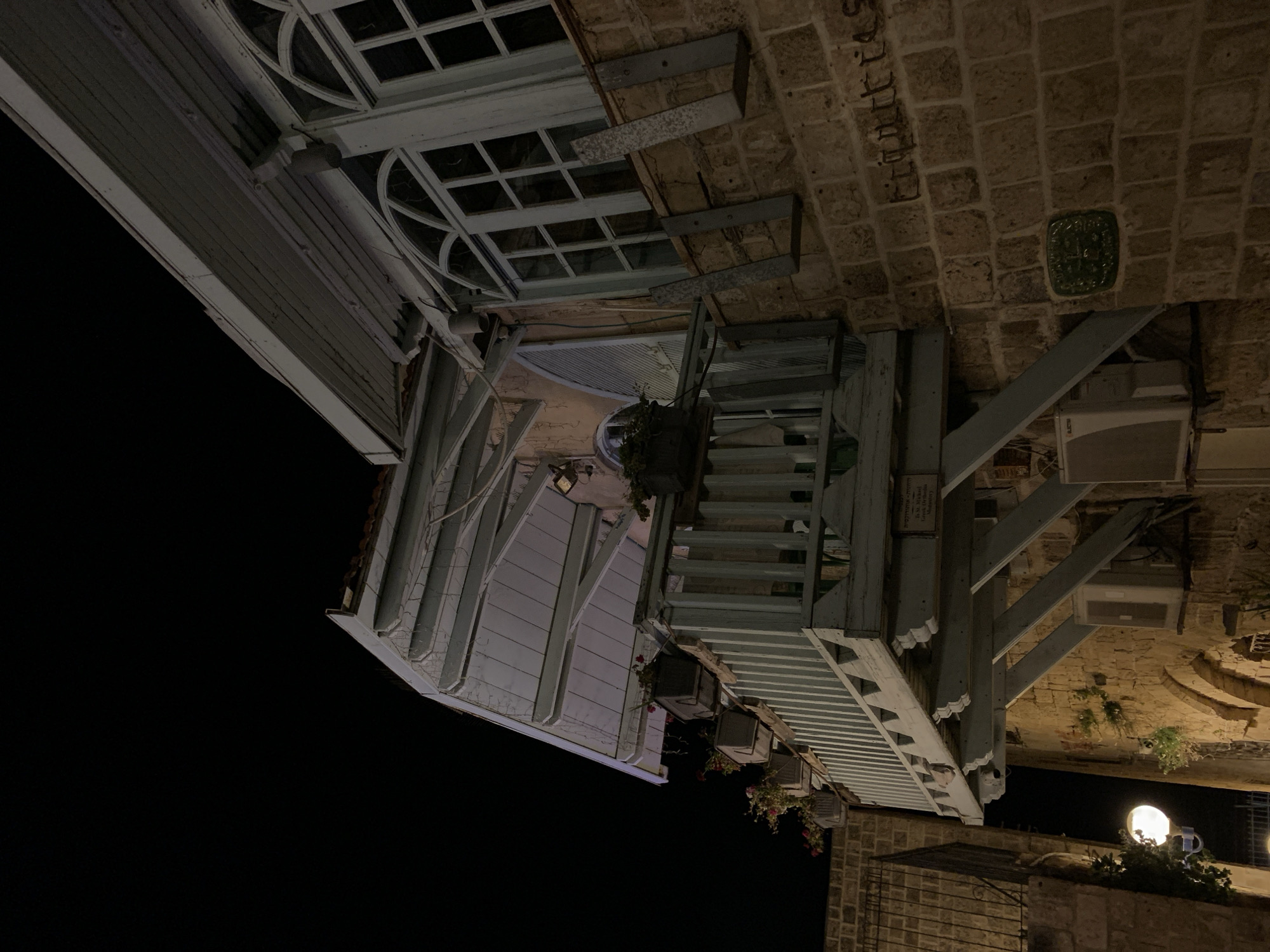 The image depicts a building in Jaffa, Tel Aviv, Israel, shot at night. The perspective is unusual, with the building appearing to lean diagonally from the upper left towards the lower right. The structure is a combination of old stonework and a more modern white wooden facade with multiple windows. Visible elements include brick walls, a wooden staircase, hanging flower pots, and an exterior lighting fixture. A patch of night sky is visible in the background.