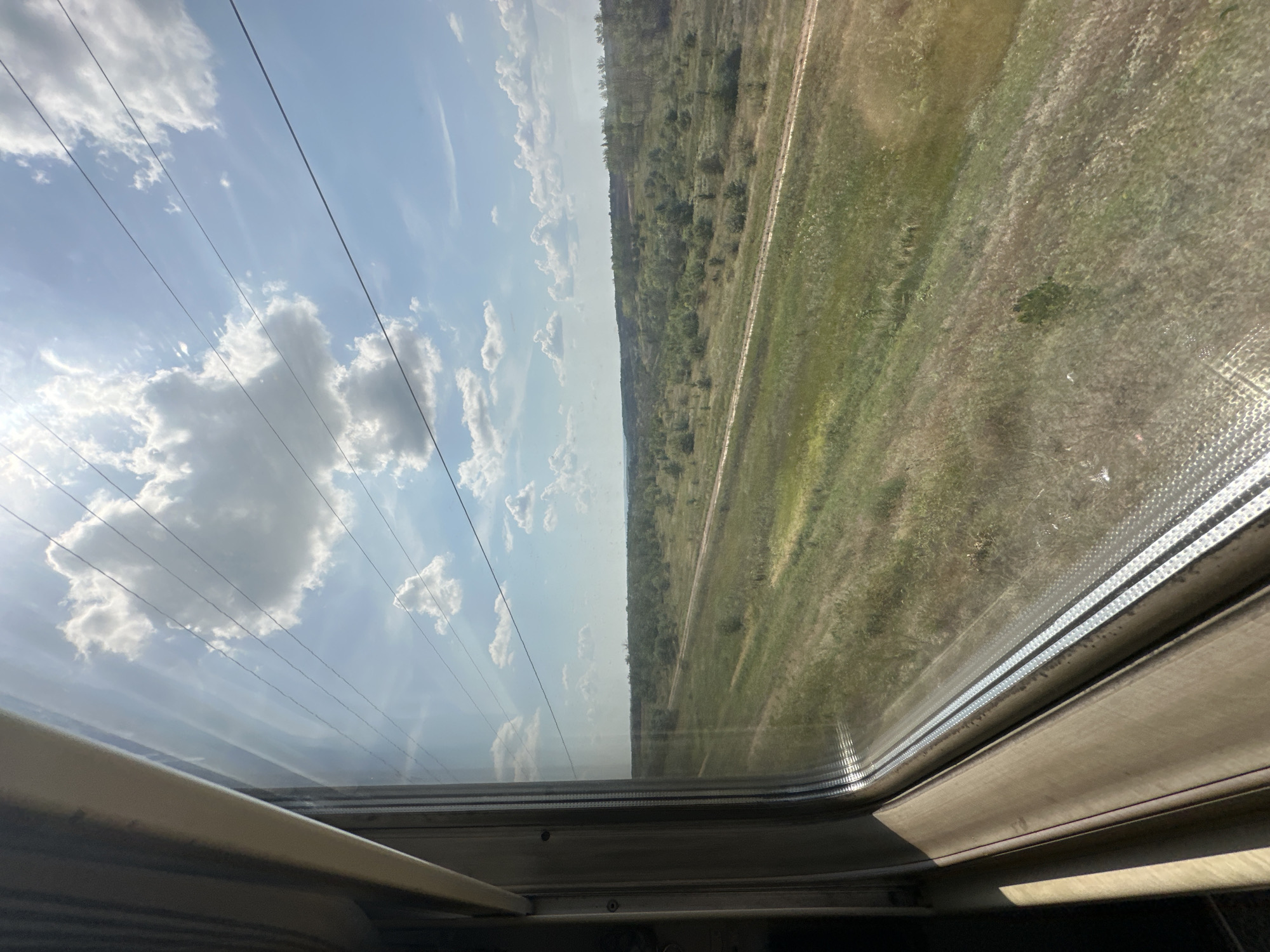 The image shows a view from a train window looking out at a rural landscape. The sky is bright blue with fluffy white clouds and a bright sun shining. There are power lines stretching across the sky. Below the sky is a vast expanse of green fields with scattered trees and bushes. A dirt road or railway track runs through the middle of the landscape. The foreground of the image shows the interior of the train, with the window frame and part of the seat visible. The overall scene depicts a journey through the countryside on a sunny day. The specific location in Kudinovka, Russia cannot be determined from the image alone.
