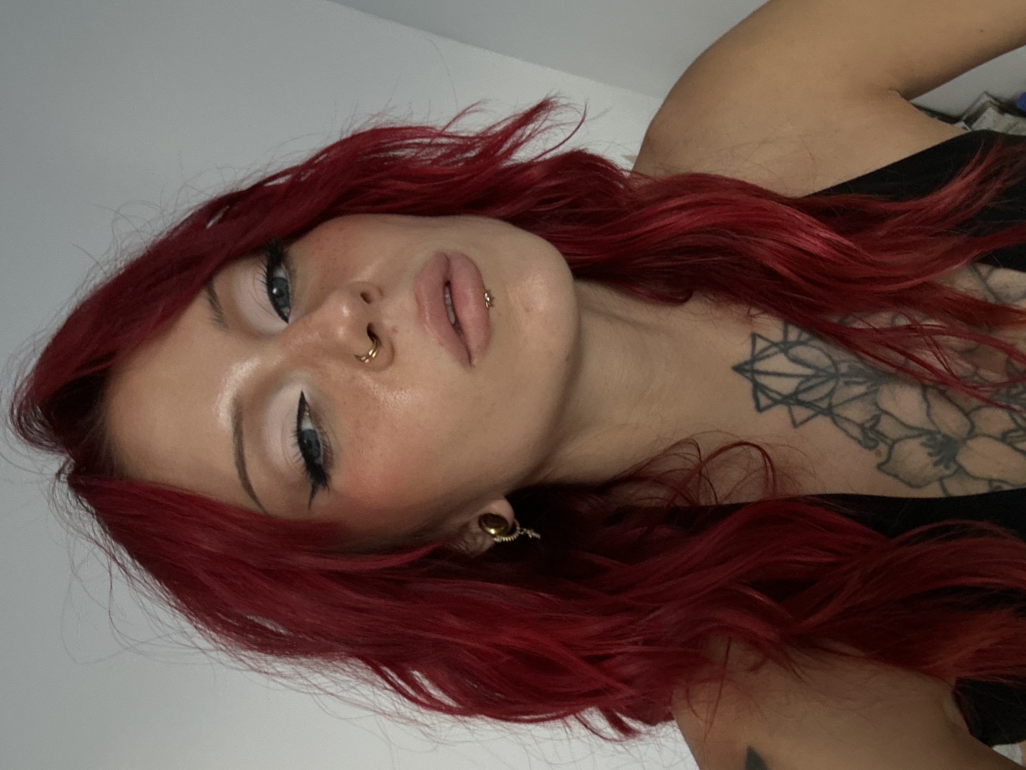 The image shows a close-up of a woman with vibrant red hair. She has striking blue eyes, prominent cheekbones, and light-colored makeup. She is wearing a nose ring and a lip ring. Her hair is styled in loose waves, framing her face. She has a tattoo on her chest and another visible on her shoulder. She is wearing a black top. The background is a plain, light-colored wall. The image does not provide any information or context related to North Kuta, Indonesia.