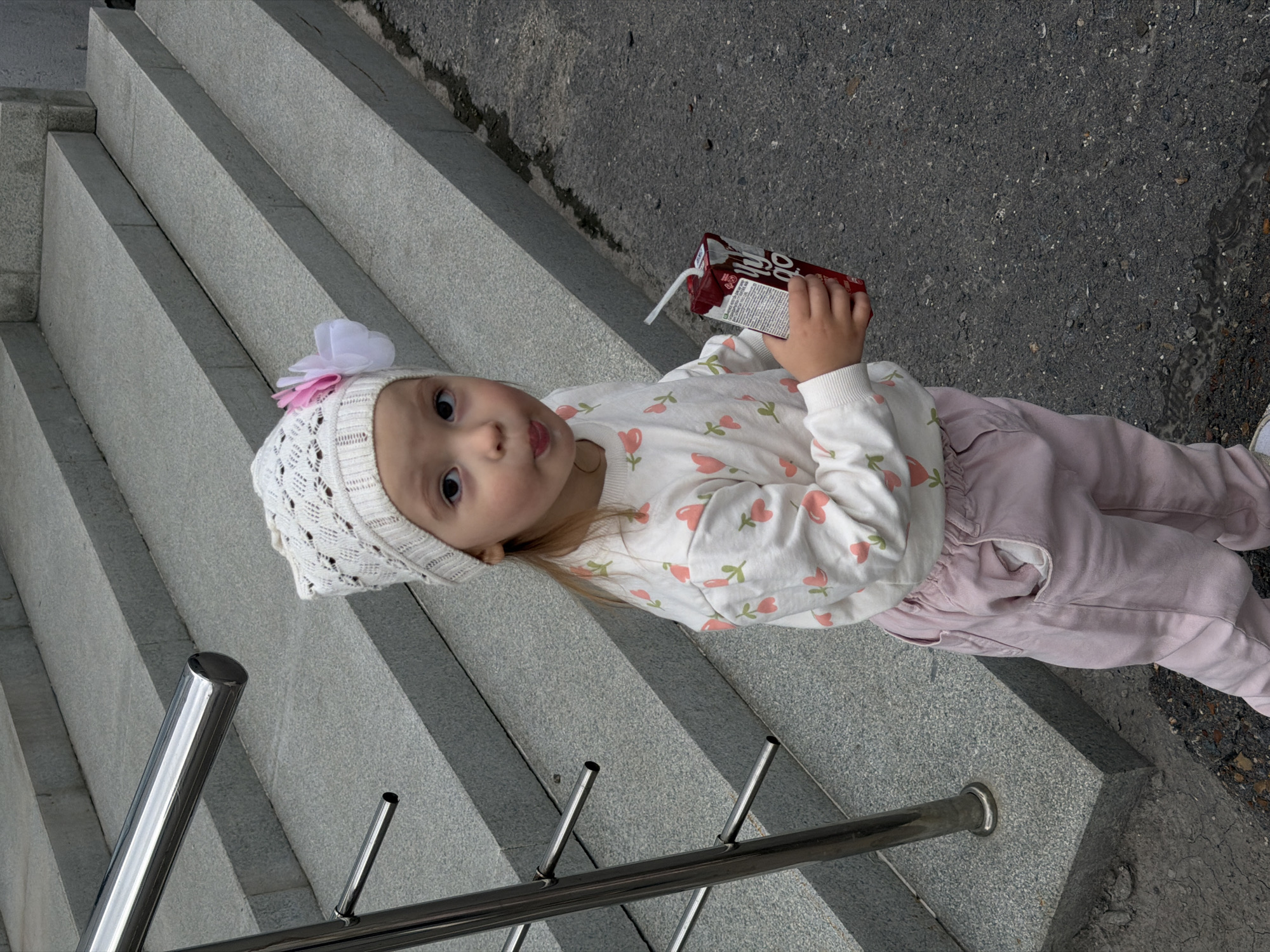 A young child is lying on a set of concrete stairs, holding a juice box with a straw. The child is wearing a white knitted hat with a pink and white flower on it, a long-sleeved white shirt with a pattern of pink flowers, and light pink pants. The child's eyes are open and looking at the camera. The child's tongue is slightly sticking out. The stairs are made of light gray concrete and appear to be outdoors, with asphalt visible in the background. There are metal railings along the stairs. The scene suggests it is daytime, and the weather appears to be overcast or it is not a sunny day. The overall mood is casual and candid. The juice box has some text on it, but it is not clearly legible. Based on the location context, the scene is in Ufa, Russia.