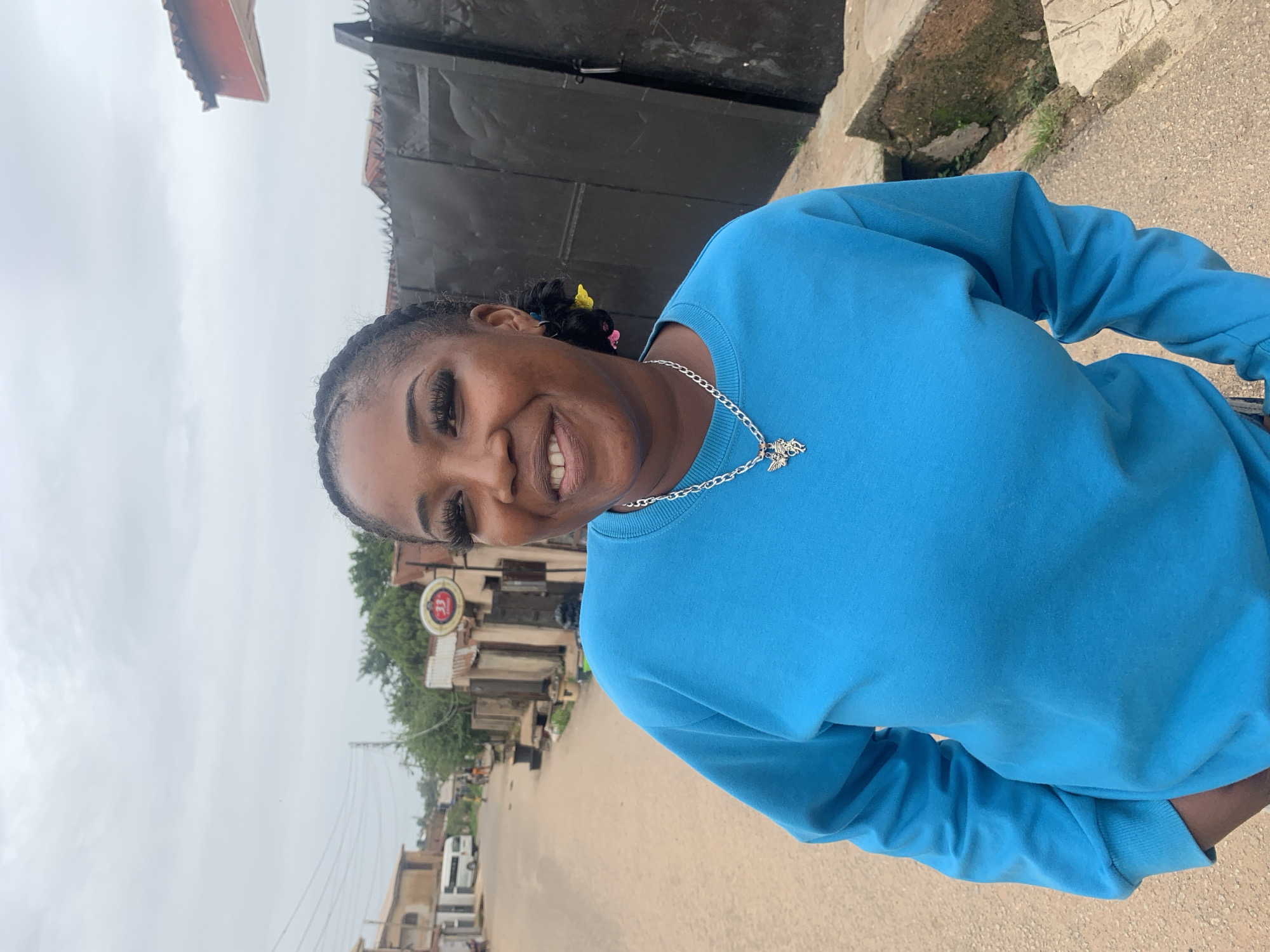 A woman is pictured in a medium shot, her body turned slightly to the right as she looks towards the camera. She is wearing a bright blue long-sleeved sweatshirt and a silver chain with a pendant. Her braided hair is pulled back, with a few colorful hair accessories visible. She has dramatic eye makeup, with long lashes. A genuine smile is on her face, conveying a friendly and approachable emotion.
The background suggests an outdoor, urban setting. A street runs through the scene, with buildings lining its sides. Some buildings appear to be made of corrugated metal or have weathered appearances. A round sign with red and white lettering, possibly a business logo, is visible on the left side of the street. Power lines crisscross the sky in the distance. The overall impression of the environment is that of a bustling, everyday scene in a town or city.
The lighting suggests it is daytime, with overcast or cloudy skies, indicated by the diffused light and the muted tones of the sky. The ground appears to be paved with concrete or asphalt, with some patches of gravel and sparse vegetation. The scene has a candid feel, as if captured in a spontaneous moment.
The visible text on the sign reads "33" in a red circle.