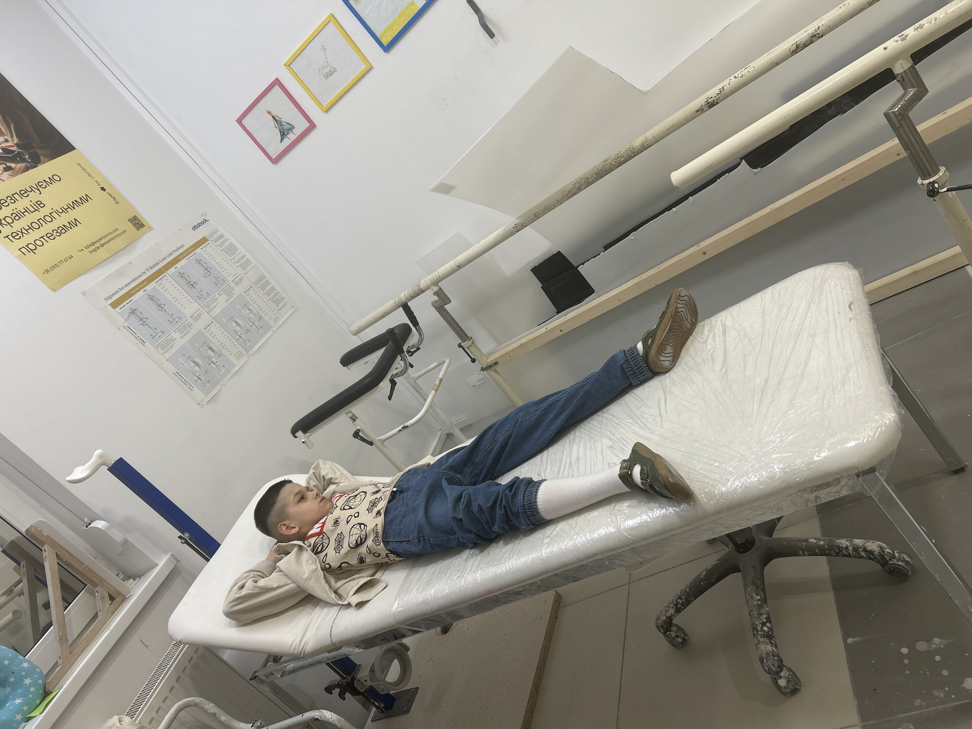 A boy is lying on a medical examination table in what appears to be a rehabilitation center or clinic. He's dressed in jeans, a white long-sleeved shirt, and brown shoes, with white socks visible at the cuffs of his jeans. The table is covered in a transparent plastic sheet.
The room has white walls and a tiled floor. Behind the table, there is a set of parallel bars, commonly used in physical therapy. A poster and some framed drawings are affixed to the wall. There is a yellow sign with the words, "безпечуємо українців технологічними протезами," and contact information. Medical equipment is seen in the background. The overall impression is a clinical setting designed for physical rehabilitation.
