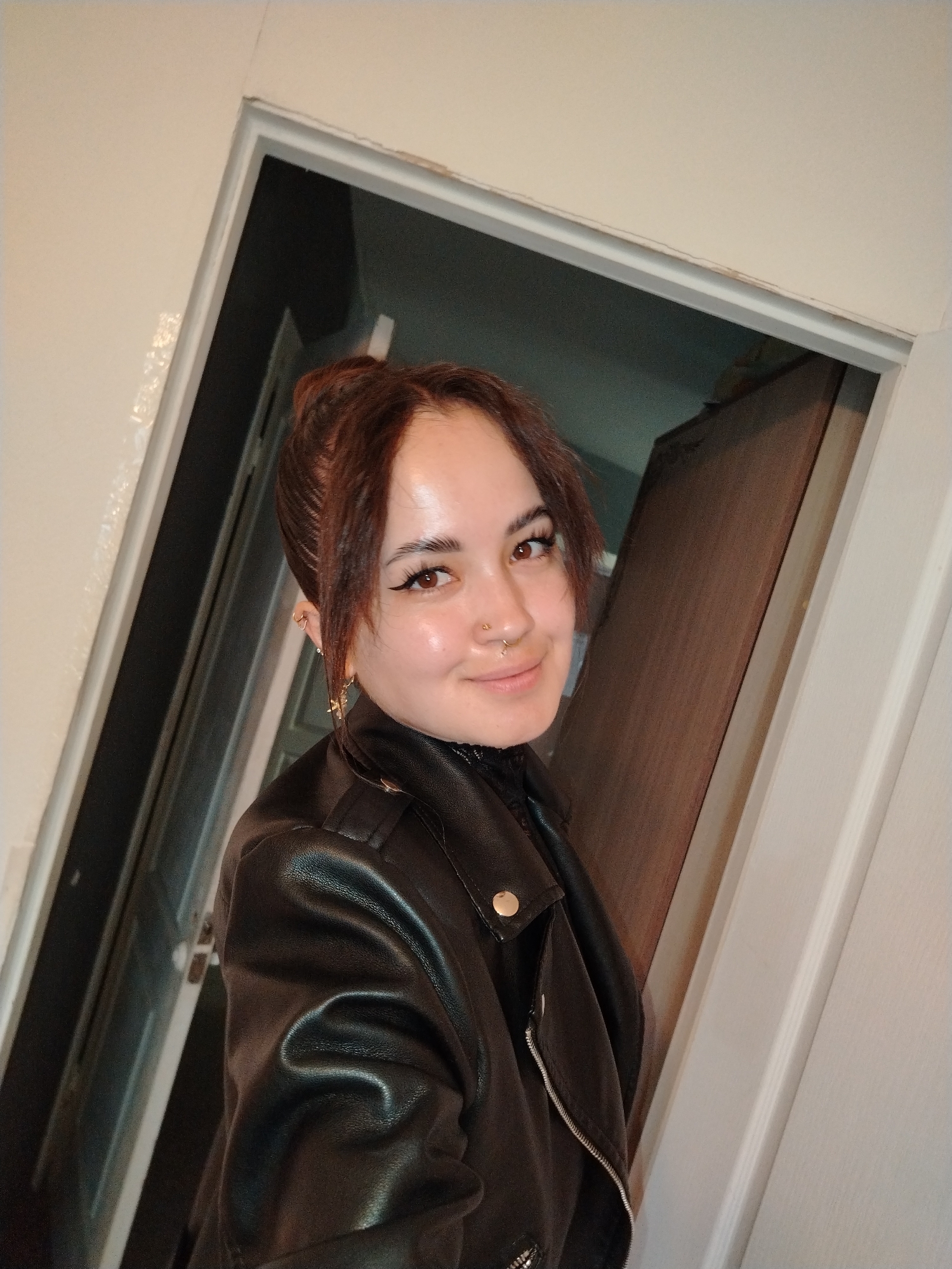 The image shows a medium shot of a young woman with auburn hair pulled up in a messy bun. She is wearing a black leather jacket with gold snaps and a black turtleneck. Her eyes are brown and she has winged eyeliner. She is also wearing hoop earrings and a nose ring. She is smiling and looking at the camera. The background appears to be a doorway with a wooden door visible. There is no visible indication of the location being Noviy, Russia.