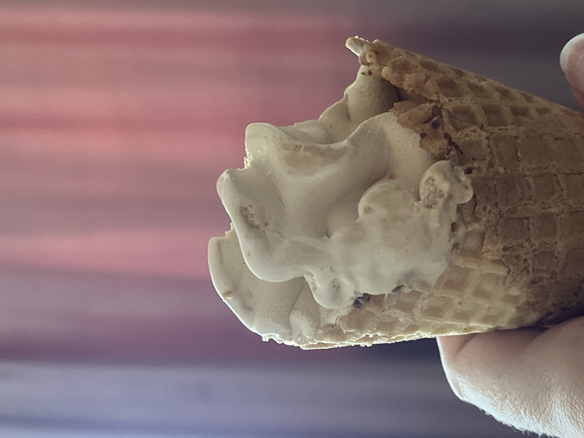 A close-up shot shows a hand holding a waffle cone filled with soft-serve ice cream. The ice cream is a light beige color and has a swirled texture, with some small dark inclusions visible, possibly nuts or chocolate chips. The waffle cone is golden brown with a grid pattern. The background consists of a blurred curtain with vertical stripes of pink and purple hues. The image appears to have been taken indoors, as suggested by the lighting and the curtain. No specific location within Krasnyye Barrikady, Russia, can be identified from the visual information provided in the image.