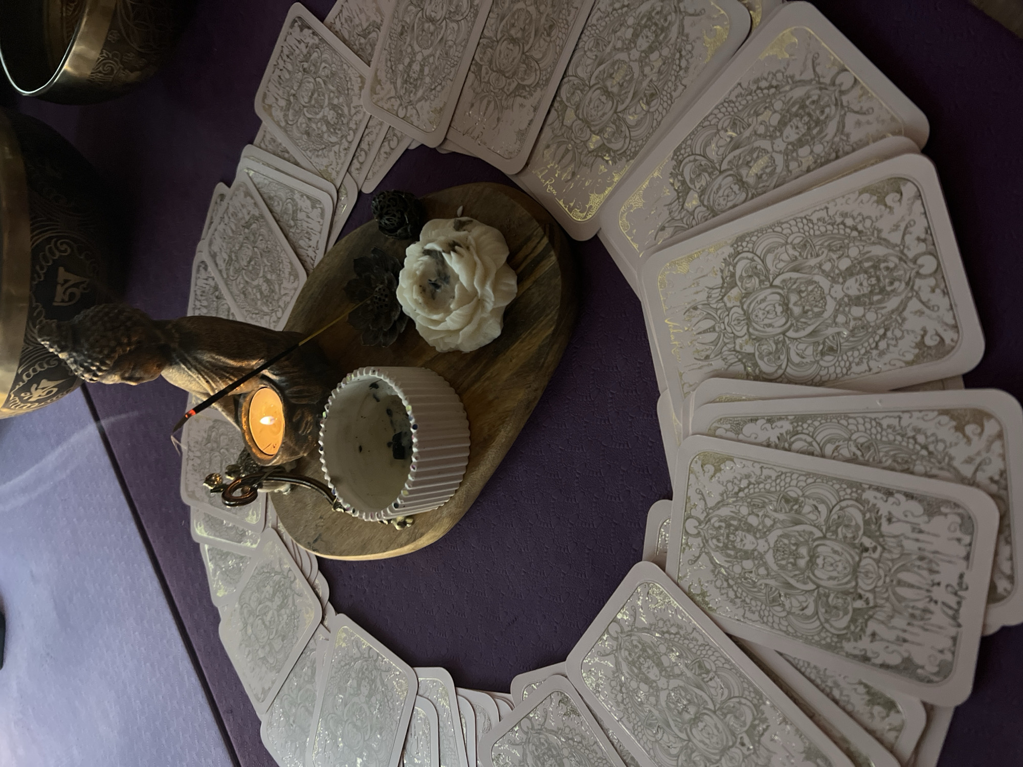 A close-up shot displays a collection of tarot cards arranged in a circular formation on a textured, purple surface. At the center of the circle lies a wooden board holding a white, floral candle, a small candle burning beside a Buddha statue, incense, and a white, ridged container. The overall setting suggests a spiritual or ritualistic atmosphere. It is not possible to determine the location of Solovej-Kljuch, Russia from this image.