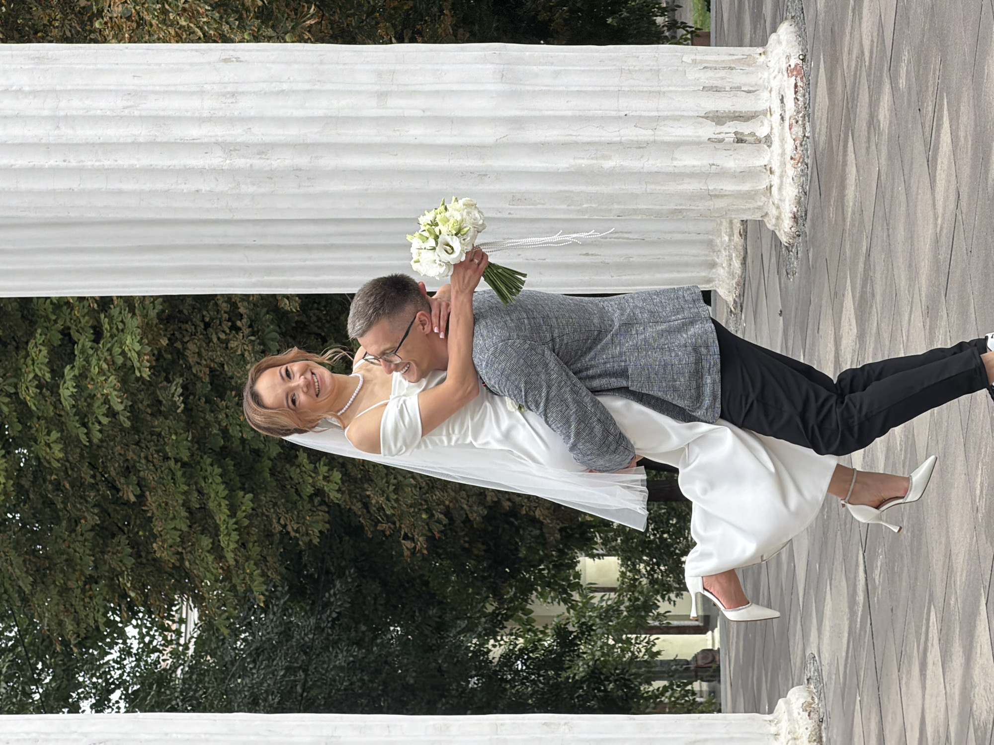 A newlywed couple is captured in a joyful moment, with the groom lifting the bride in his arms. The bride, wearing a white off-the-shoulder wedding dress and a veil, is smiling at the camera. She is holding a bouquet of white flowers. The groom, dressed in a grey checkered jacket and black trousers, is looking at the bride. They are standing in front of large, weathered white columns, likely part of an architectural landmark. The background shows lush green trees. The scene appears to be outdoors, on a paved surface made of square tiles. The image is likely taken in Dnipro, Ukraine, given the style of architecture and the presence of classical columns often found in public spaces and parks within the city.