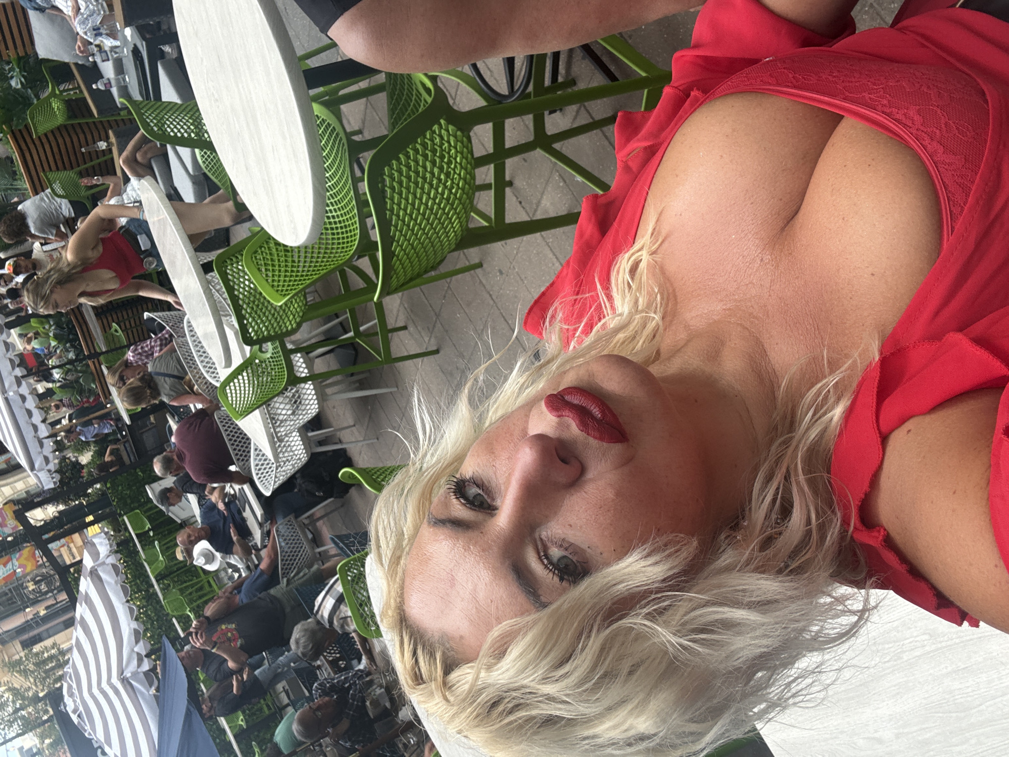 The image depicts an outdoor seating area at a restaurant or bar, likely in an urban setting. The foreground features a woman with blonde hair and red lipstick, wearing a red top that reveals her cleavage. The background shows several tables and chairs, some occupied by people. There are also umbrellas providing shade. The overall atmosphere appears casual and social. While the image is stated to be from Calgary, Canada, there are no specific visual cues within the image to confirm this location, such as recognizable landmarks or signage. The scene is likely set during daytime, judging by the lighting.