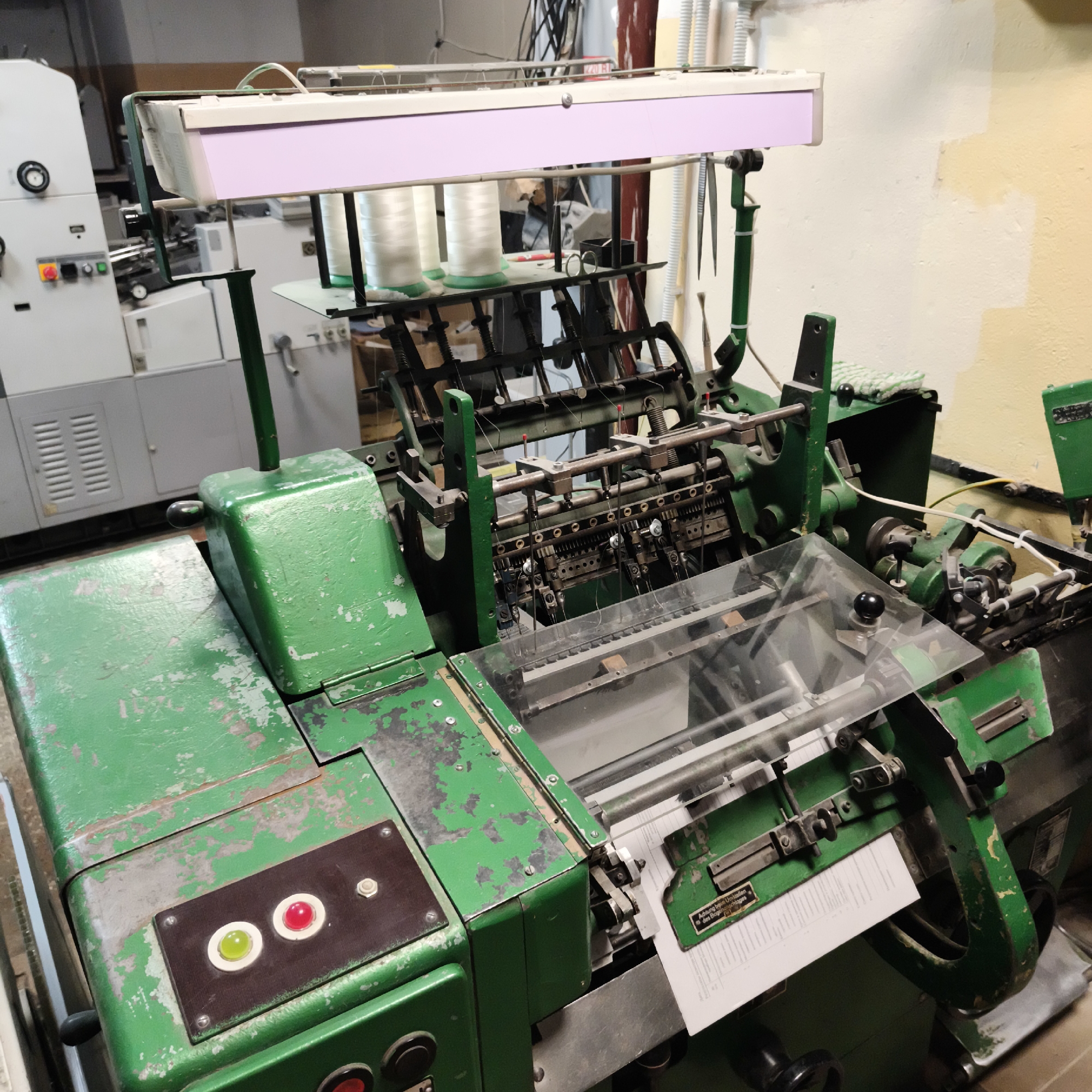 The image shows a close-up view of an industrial sewing machine, likely in a workshop or factory setting, located in Chaikovsky, Russia. The machine is predominantly green, with signs of wear and tear, including chipped paint. It features multiple spools of white thread, needles, and various mechanical components, suggesting it is a specialized piece of equipment for mass production. A clear plastic guard covers part of the mechanism. To the left, a portion of another piece of machinery, possibly a printer or labeler, is visible, also appearing to be industrial. The background includes a wall that is partly painted a light cream color and partly a yellow-ochre hue, with some lighter patches suggesting previous paintwork. The overall impression is one of an operational but older industrial environment. There are no people visible in the image.