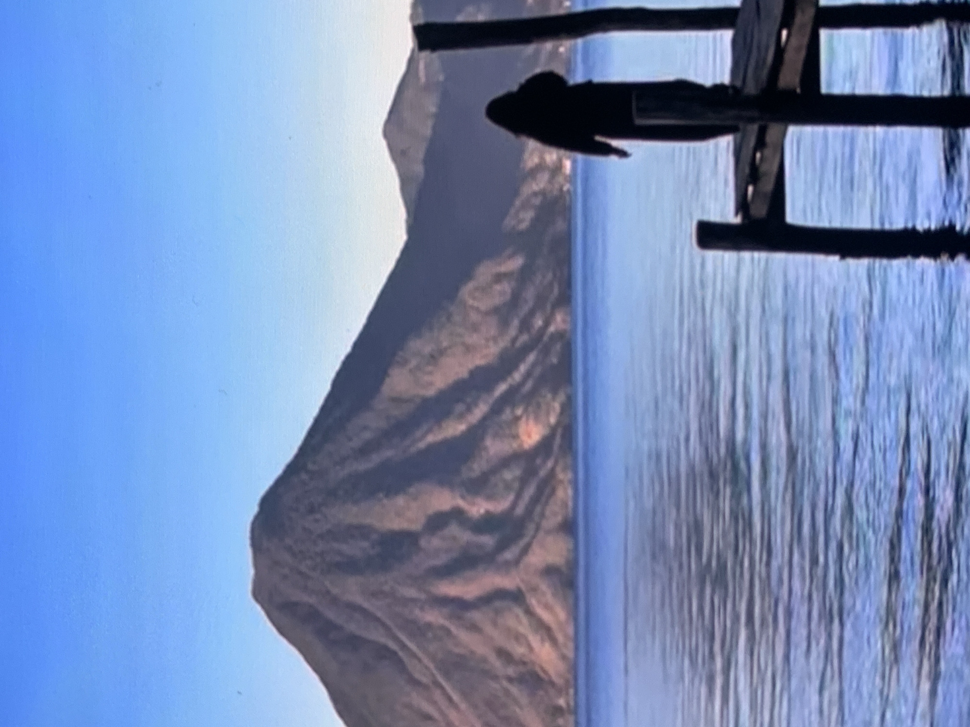 A silhouette of a person wearing dark clothing stands on a wooden dock. The person is facing away from the camera, looking out at a body of water. In the background, a large, dormant volcano rises above the water, with smaller mountains behind it. The sky is clear and blue. The scene is tranquil and serene. This image is not located in Stavropol, Russia.