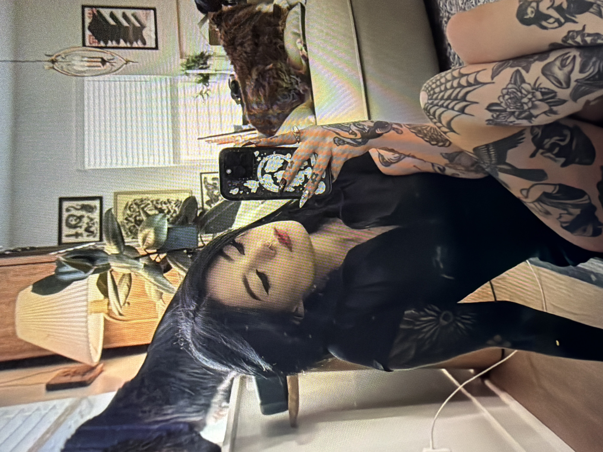 In the foreground, a woman with dark hair and dramatic eye makeup takes a selfie in a room. She is wearing a black top and has extensive tattoos on her arms and legs, including a spiderweb on her knee and floral designs. She is holding a phone with a butterfly-themed case. Behind her, a large black dog with its back to the camera is perched on a windowsill, and a fluffy brown dog is resting on a sofa. The room appears to be a living area with framed artwork on the walls and a floor lamp. The scene is indoors, and there is no indication of the city of Shumen, Bulgaria.