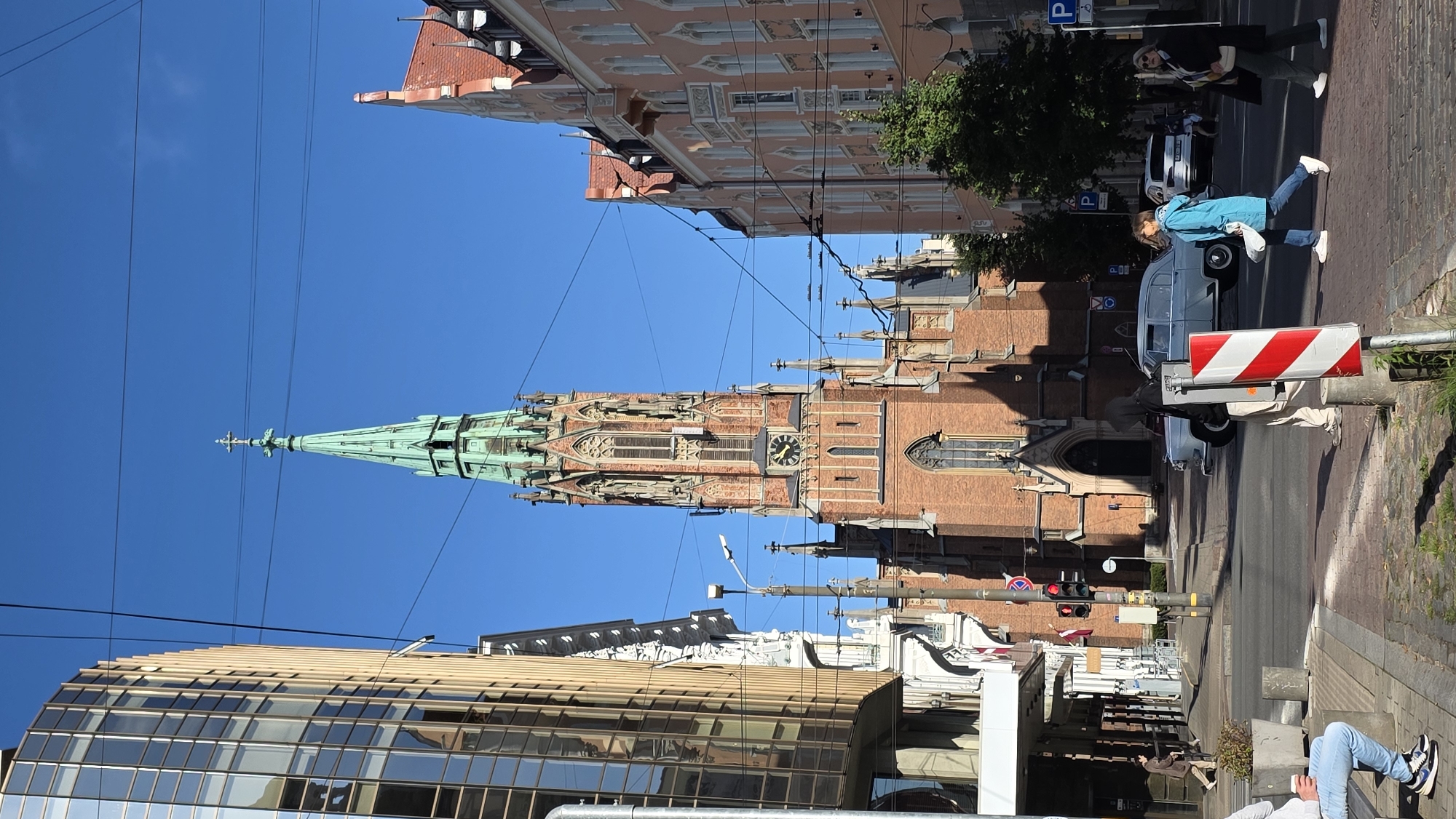 A street scene in Riga, Latvia, features a tall, ornate brick church with a green spire as the central focus. The church has a clock visible on its facade. To the left of the church is a modern glass-fronted building, and to the right, a pink-toned building with decorative balconies lines the street. Power lines crisscross the sky above. Several people are visible on the sidewalk and crossing the street. A vintage grey car is parked or driving in the street, and a white modern SUV is parked further down. Pedestrians are walking and crossing the street. The weather appears sunny with a clear blue sky. The foreground shows a paved sidewalk with a red and white striped post.