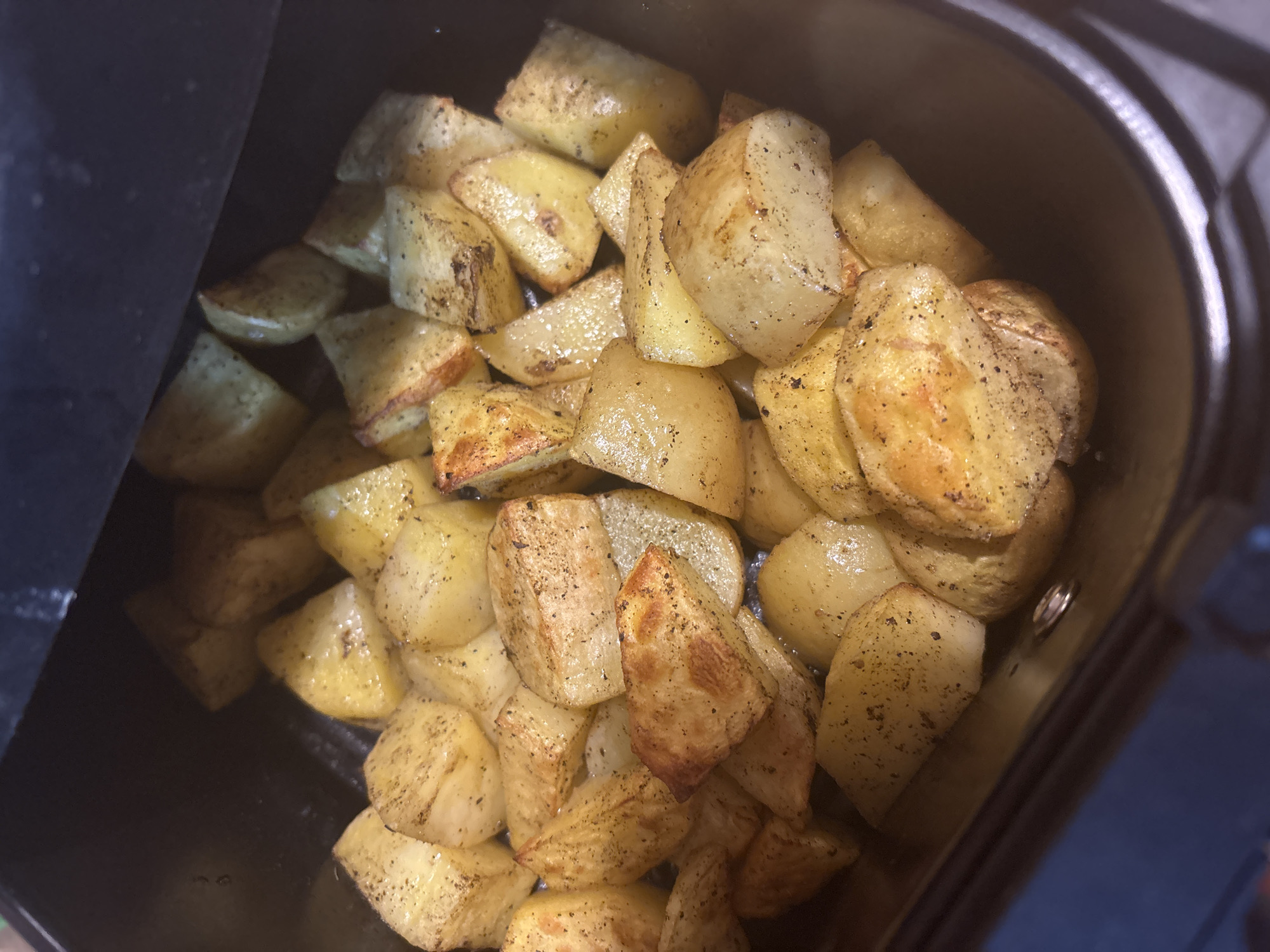 The image shows a close-up of golden-brown, cooked potato chunks inside a dark-colored appliance, likely an air fryer. The potatoes appear to be seasoned with black pepper and possibly other spices. The potatoes are arranged densely within the fryer basket. The appliance's interior is black or dark gray, and the edges of the appliance are visible. The image provides no visual information that allows it to be located in Sofia, Bulgaria.
