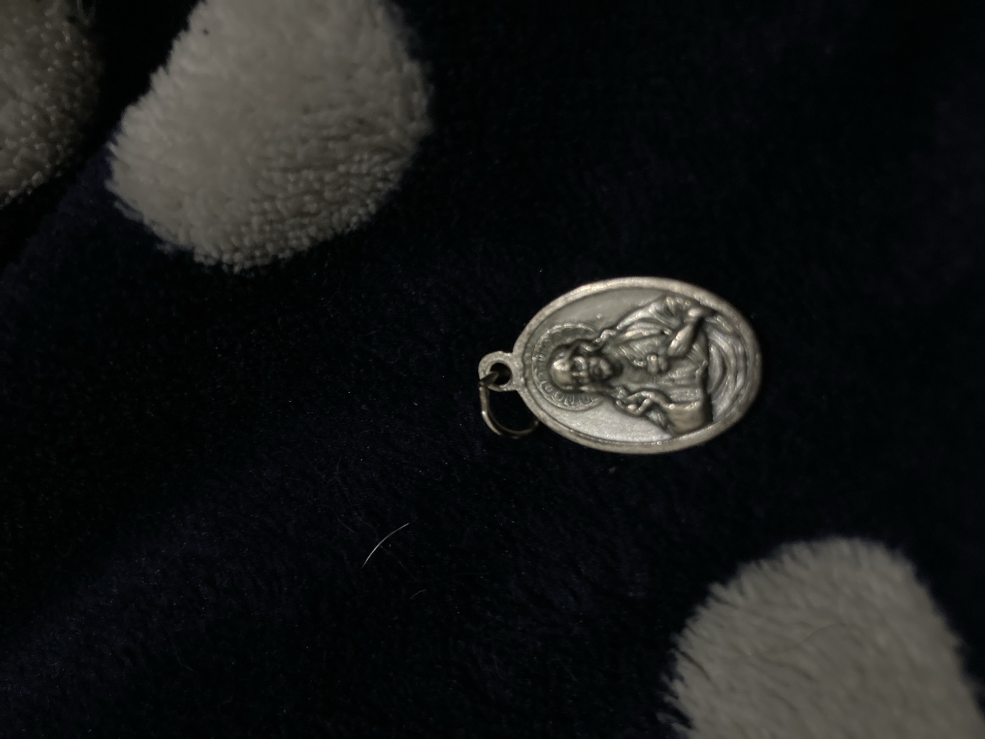 A close-up photograph displays a silver-colored, oval-shaped medallion resting on a dark, plush, and fuzzy fabric, possibly a blanket or garment. The fabric features several lighter, circular or amorphous textured patterns.
The medallion is embossed with the detailed depiction of a male figure, shown in profile facing to the right. The figure has a beard and appears to be wearing robes. A halo or stylized aura is visible around the figure's head. The left arm of the figure is visible, seemingly making a gesture or holding an indistinct object. The iconography strongly suggests a religious figure, possibly Saint Joseph.
Faint, unreadable text is visible around the perimeter of the medallion, particularly near the figure's head. A single letter, possibly "S.", can be partially discerned at the top left edge. The medallion has a small metal loop at its top, indicating its use as a charm or pendant.
The photograph is a still-life, taken in dim lighting conditions, suggesting an indoor setting. No specific activity or event is taking place. A thin, light-colored strand or fiber lies on the dark fabric beneath the medallion.