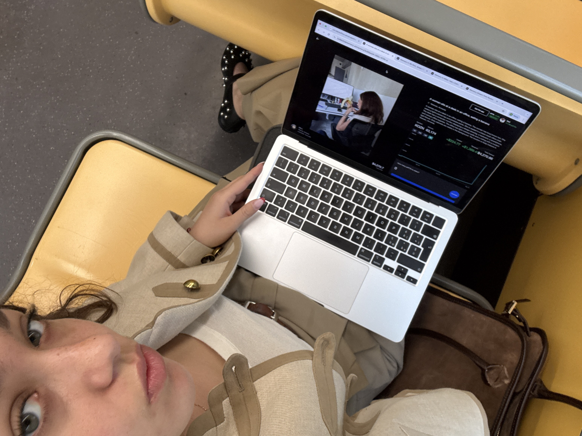 A high-angle selfie captures a woman seated in a public transport vehicle, likely a train or bus in Milan, Italy, as indicated by the yellow-toned seats and grey speckled floor. She looks directly at the camera with a neutral expression. She wears a light-colored, possibly linen, jacket or blazer with gold buttons over a white top, and tan trousers. Her left hand is positioned on the keyboard of an open silver MacBook Pro, which rests on her lap.
To her left, a brown leather bag or briefcase is visible on the seat. In the background, her left foot, adorned in a black flat shoe covered in silver studs, is visible. The laptop screen displays a web browser with multiple tabs. The main content shows a video frame of a woman at a desk in an office, holding a banana. Below the video, text reads "A woman sit at a desk in an office, eating a banana". Further down, there's financial information, including "KOZN 63.174" and monetary figures such as "+$224.77 +$1,384 $4,270.66", alongside a graph, suggesting a stock market or cryptocurrency interface. The QWERTY keyboard layout is visible, with keys including W, E, R, T, Y, U, O, P, S, D, F, G, H, J, K, L, Z, X, C, V, B, N, M. The scene depicts a person engaged with their laptop while commuting.