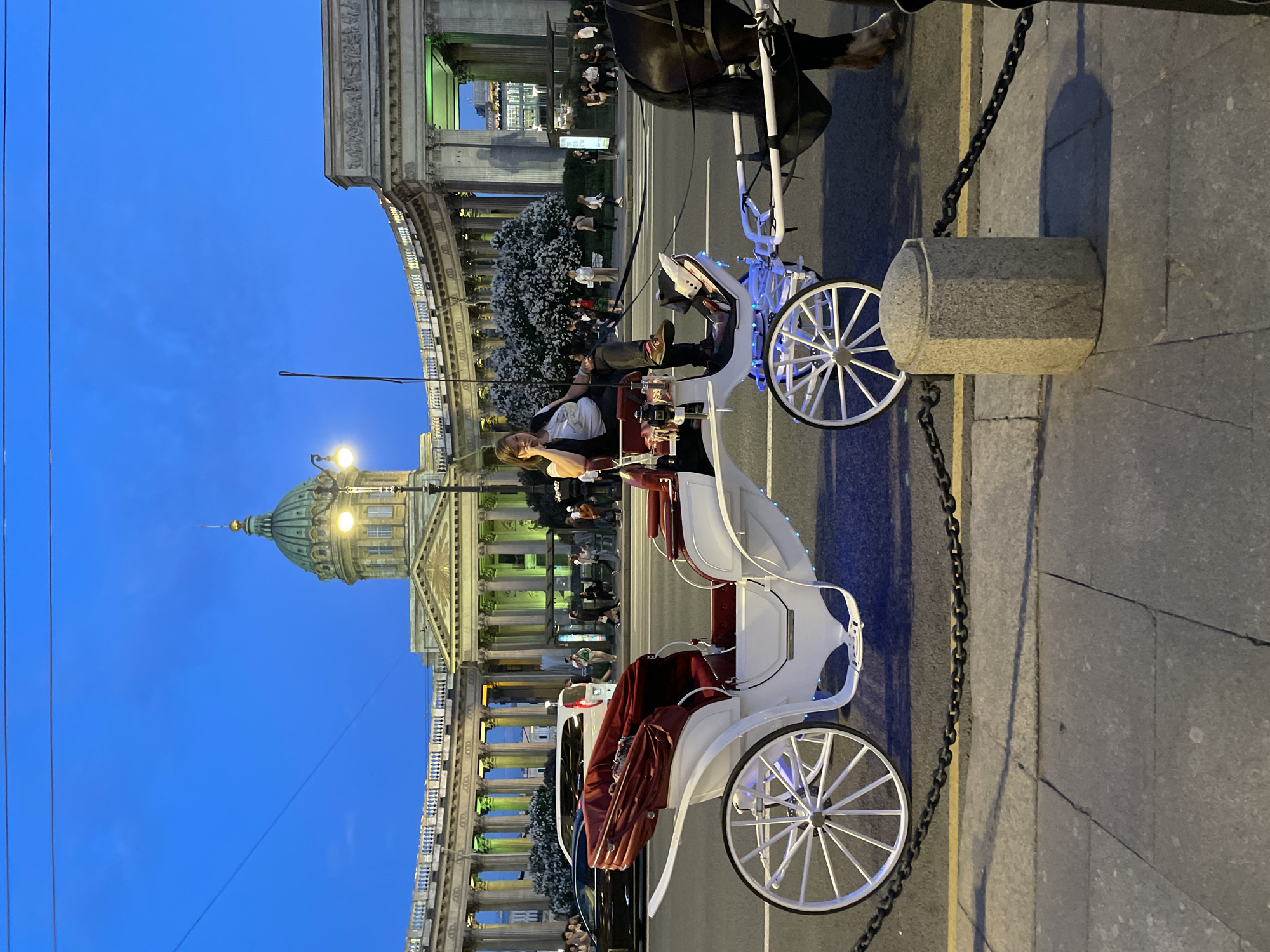 The image captures an early evening scene on Nevsky Prospekt in Saint Petersburg, Russia, with the Kazan Cathedral dominating the background under a deep blue twilight sky. A white horse-drawn carriage with red upholstered seats is positioned in the mid-ground, adjacent to the sidewalk. A woman with light-colored hair, wearing a white t-shirt, dark vest, and dark pants, is seated in the carriage, looking towards the left with her right hand resting on her cheek. The head and upper foreleg of a dark horse are visible on the far right, harnessed to the carriage. The carriage features white spoked wheels and small blue accent lights along its lower frame.
The Kazan Cathedral in the background showcases its green dome and a prominent semi-circular colonnade, illuminated with a mix of green and yellow lights that highlight its architectural features. A bright streetlight is visible near the cathedral's dome. The street between the carriage and the cathedral is dark asphalt, with several vehicles visible, including a white van and a dark sedan. A line of trees or bushes with white blossoms separates the street from the cathedral's immediate vicinity. Numerous pedestrians are distributed along the sidewalks and in front of the cathedral in the background.
In the immediate foreground, a black metal chain extends along the edge of the paved sidewalk, connected on the right to a light-colored, octagonal stone bollard. Overhead utility wires are faintly visible against the blue sky. The scene is illuminated by artificial light sources, contrasting with the natural twilight.