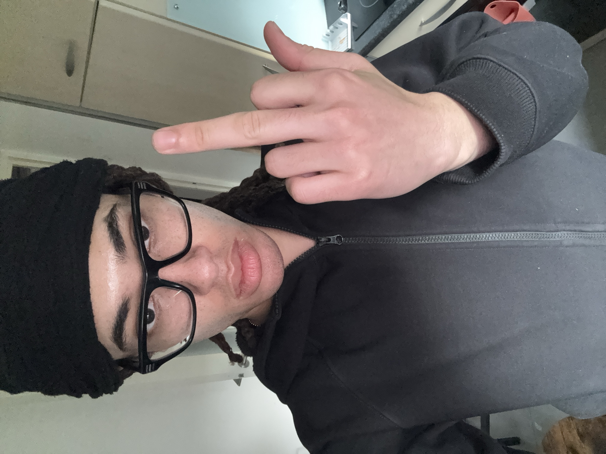 A person is depicted in a close-up, rotated indoor photograph. The individual, with a light skin tone, wears a black knitted beanie, large black-rimmed glasses, and a black zippered hoodie or jacket. Dark braided hair is visible beneath the beanie. The person's face is central to the frame, with their eyes looking directly at the camera and lips slightly parted.
Their right hand is raised, making a gesture with the middle finger extended towards the viewer. The background features light-colored cabinets above and a darker countertop with what appears to be a stove burner or appliance visible. A hint of a red object, possibly fabric, is in the upper right corner of the background. The setting suggests a domestic environment, likely a kitchen or a room with kitchen elements. The lighting appears to be consistent indoor illumination. No text is visible in the image.