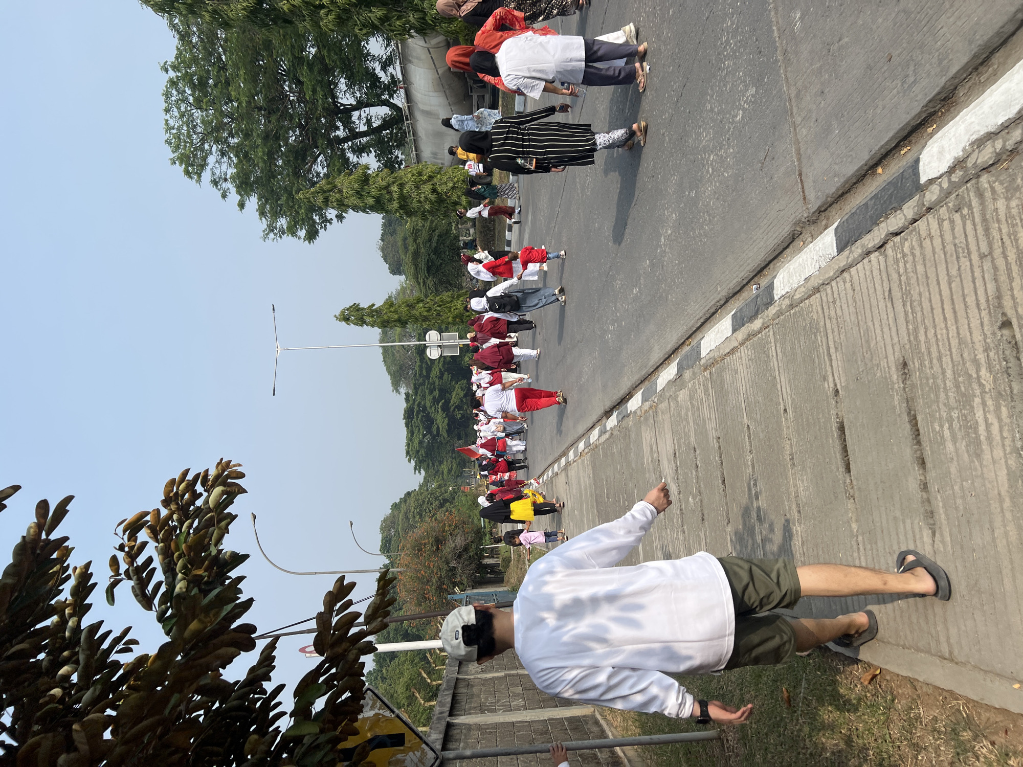In Klapanunggal, Indonesia, a group of people are walking on a road and a sidewalk. The person in the foreground is wearing a white long-sleeved shirt and olive green shorts, walking on the sidewalk away from the viewer. They are also wearing a white cap and black flip-flops. Behind them, on the road, a procession of people are walking, many of whom are wearing red and white attire, consistent with Indonesian national colors. Some individuals are carrying Indonesian flags. The environment includes a concrete wall on the left side of the sidewalk, trees and foliage lining the road, and streetlights. The sky is clear and blue.